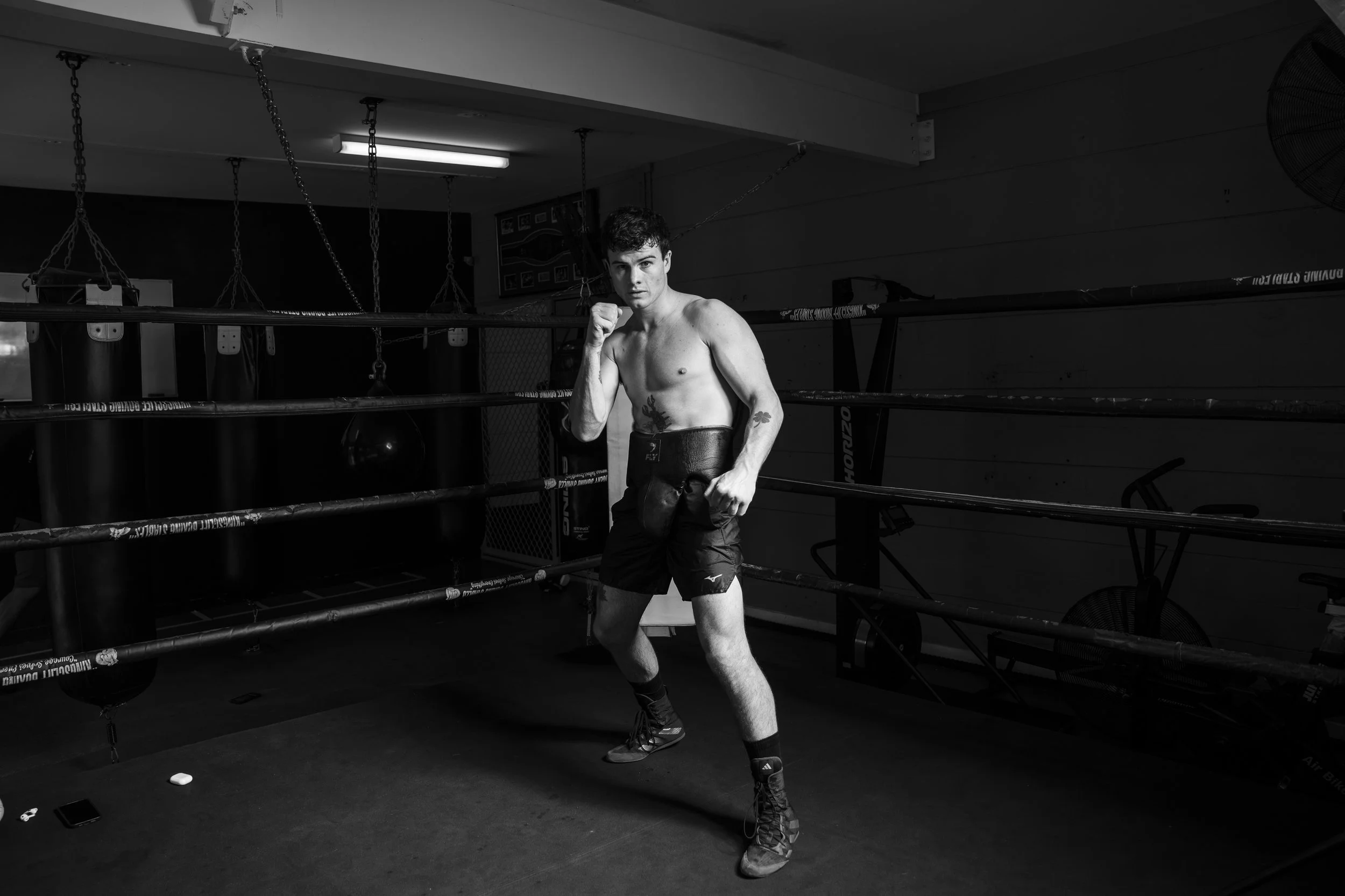 Black and white image of a shirtless man boxing in a gym ring, wearing shorts and boxing shoes, in a fighting stance. Gold Coast | Byron Bay Photography by Joey Bailey