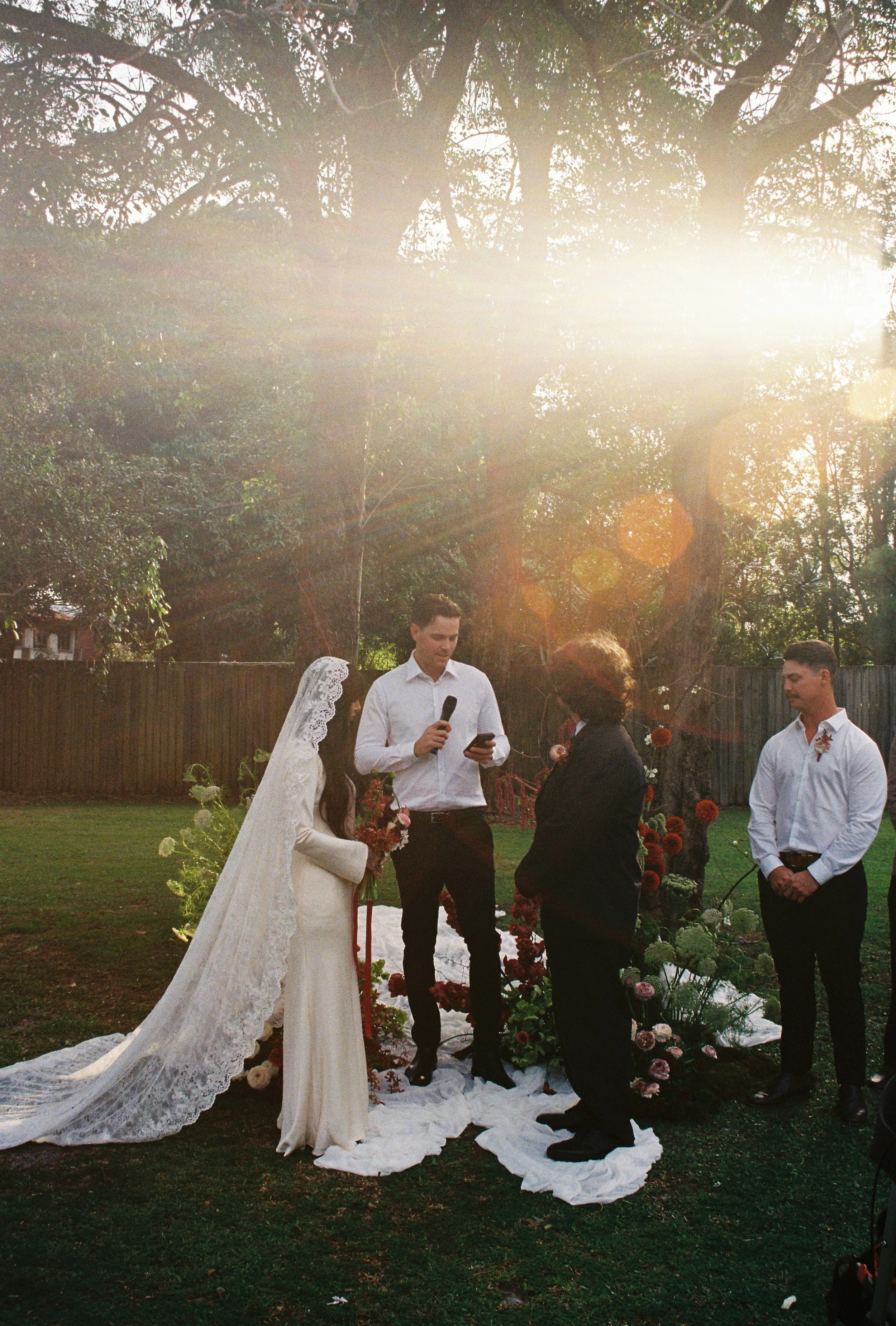 A wedding ceremony taking place outdoors during sunset, with the bride in a long white dress and veil, and the groom in a white shirt and dark pants, facing each other in front of a floral altar surrounded by floral arrangements and greenery.