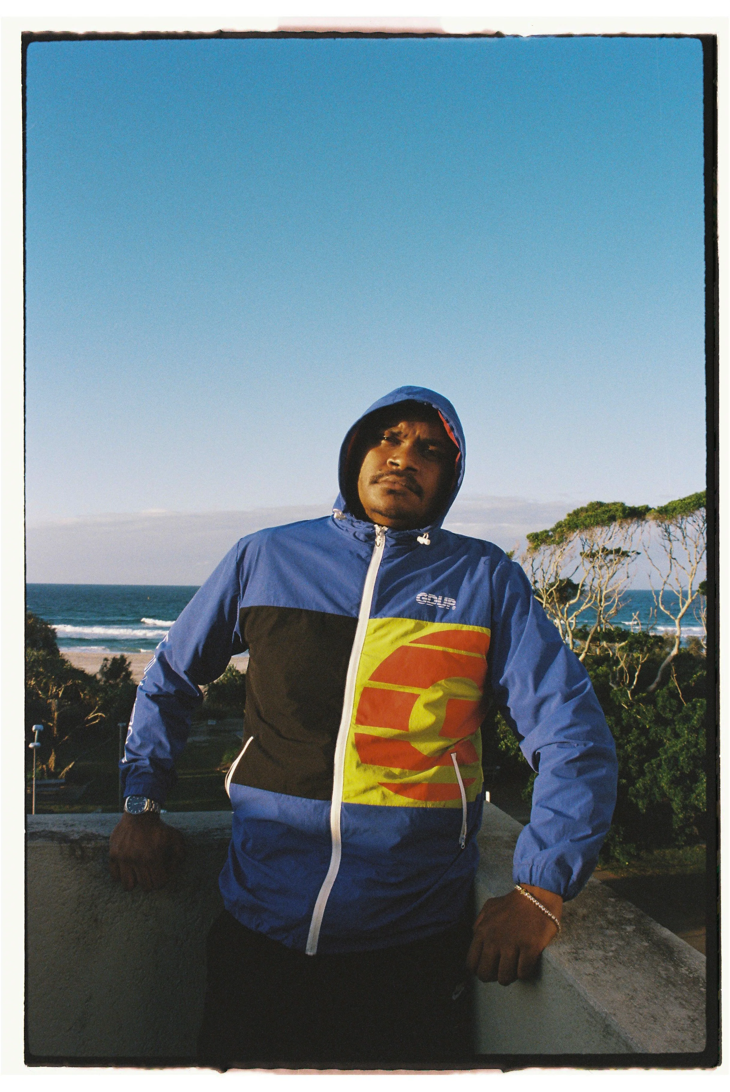 Nate G standing outdoors on a sunny day, wearing a blue and black jacket with a yellow and red abstract design, with a background of trees, ocean, and clear sky. Photography by Joey Bailey
