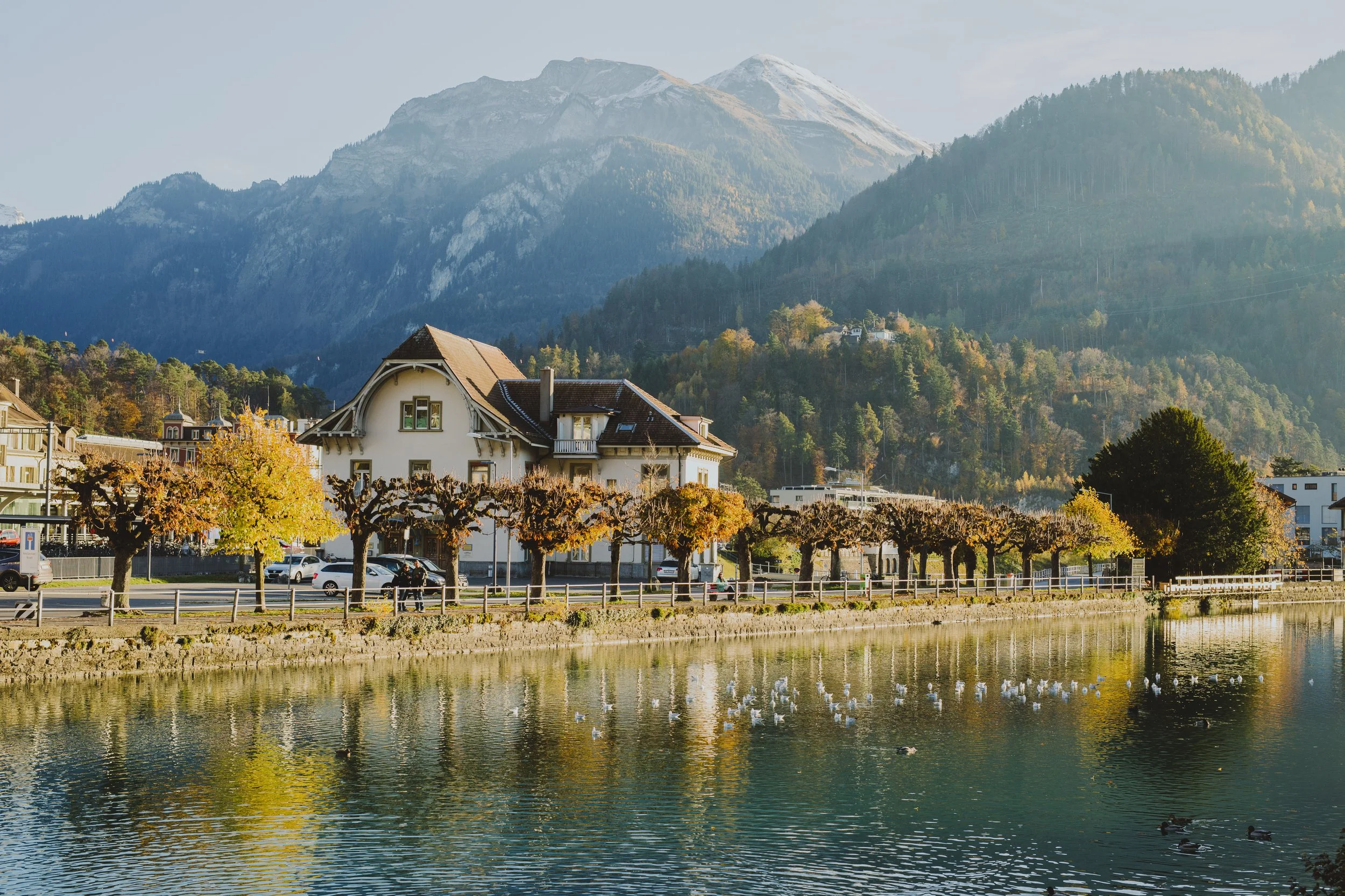 Scenic view of a building near a lake with autumn trees and mountains in the background.