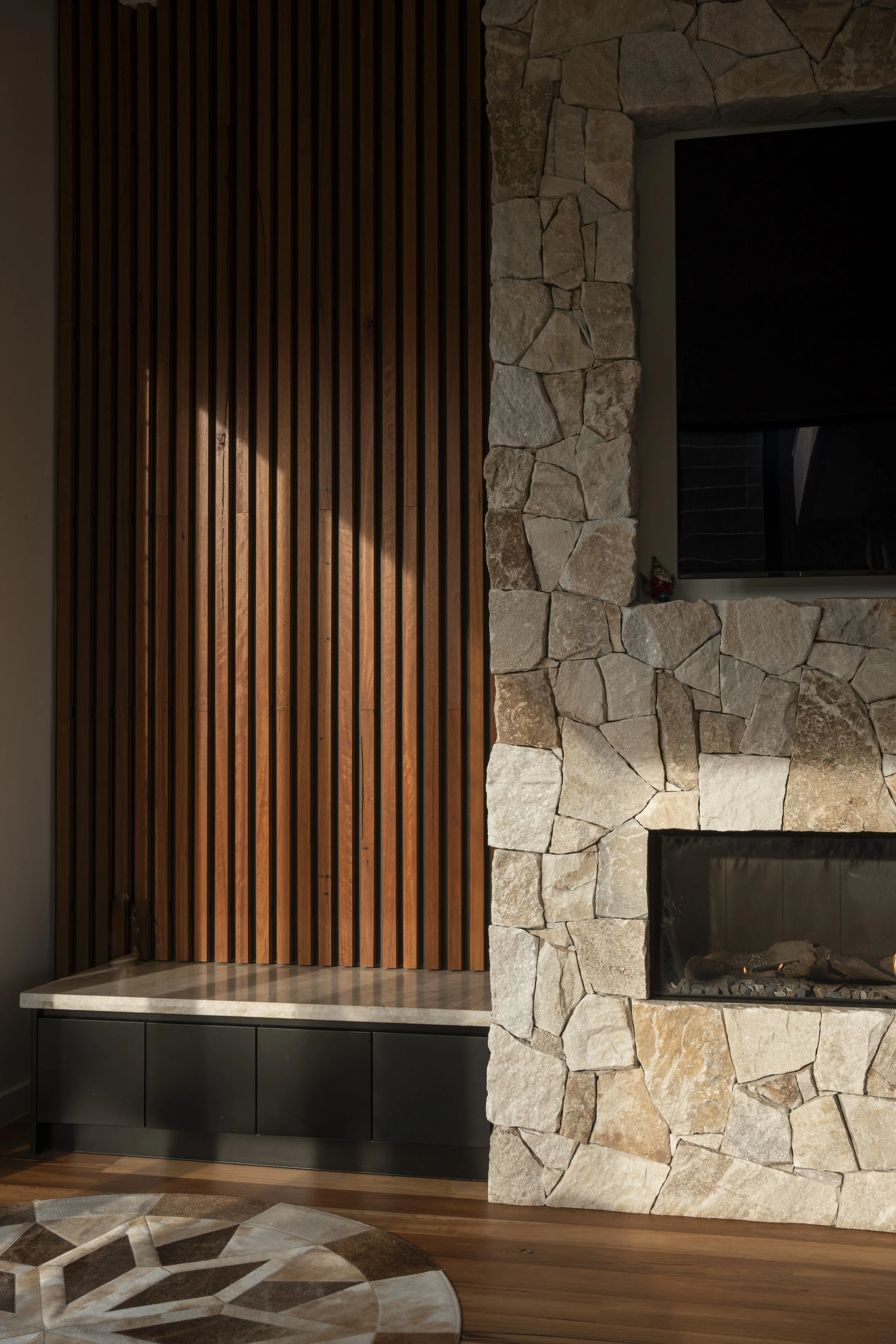 Interior living room with wooden slat wall, stone fireplace, television, and a patterned round rug on hardwood floor. Architecture photography by Joey Bailey Gold Coast