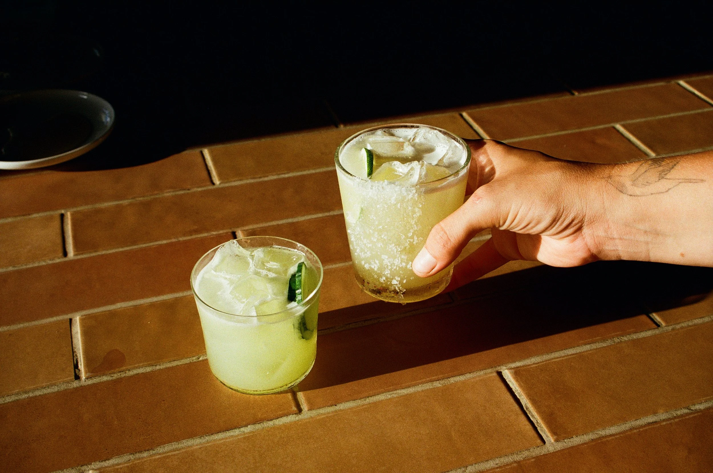 Two glasses of margaritas with ice and lime slices on a wooden table, one being held by a hand.Gold Coast | Byron Bay Food Photography by Joey Bailey