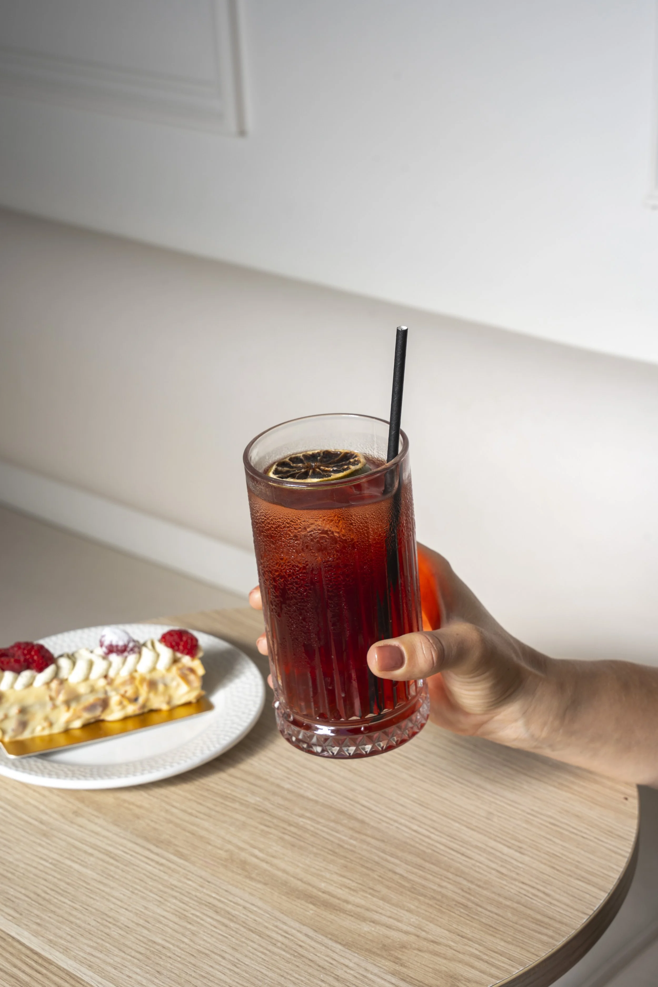 Hand holding a glass of iced red beverage with a straw and lemon slice, next to a dessert topped with whipped cream and raspberries on a plate, on a wooden table. Gold Coast | Byron Bay Food Photography by Joey Bailey