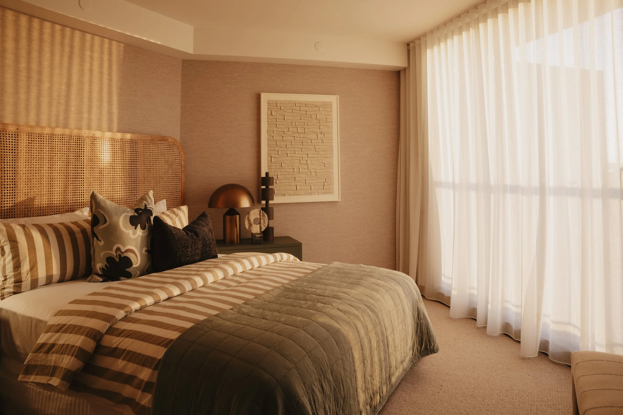 A bedroom with a neatly made bed, patterned pillows, a bedside table with a brass lamp, a framed piece of art on the wall, and large sheer curtains letting in natural light. Gold coast architecture photographer