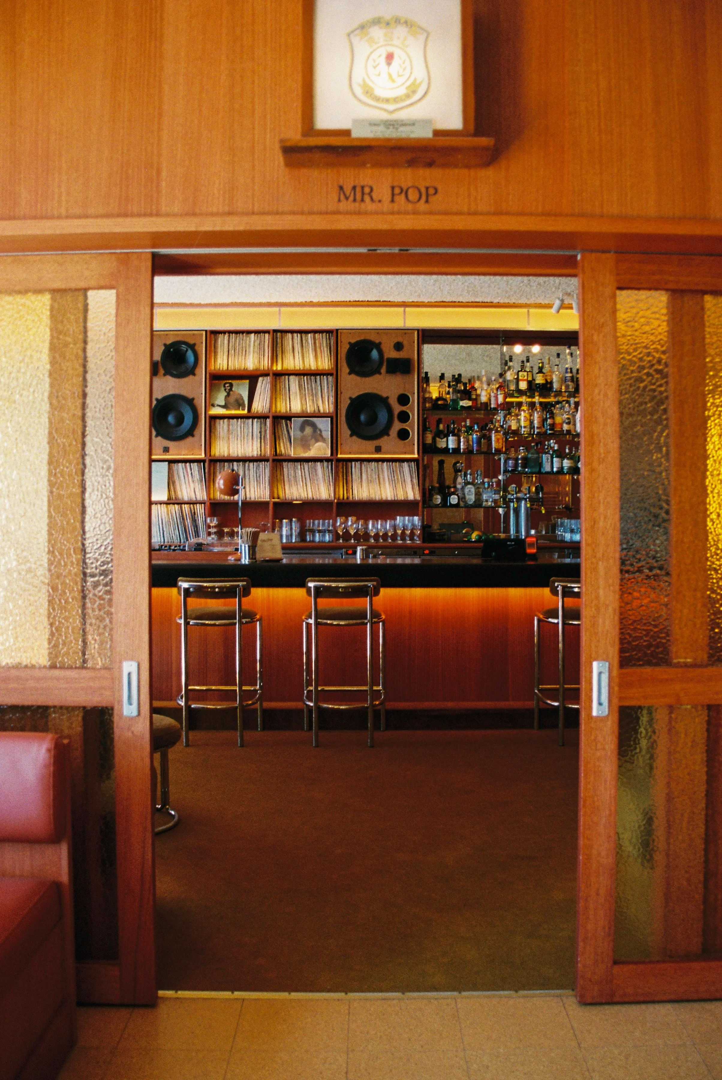 A view of a bar within a restaurant, seen through a wooden sliding door. The bar has three metal chairs with dark cushioned seats, a black countertop, and shelves filled with liquor bottles. Behind the bar, there are audio speakers and a vinyl record