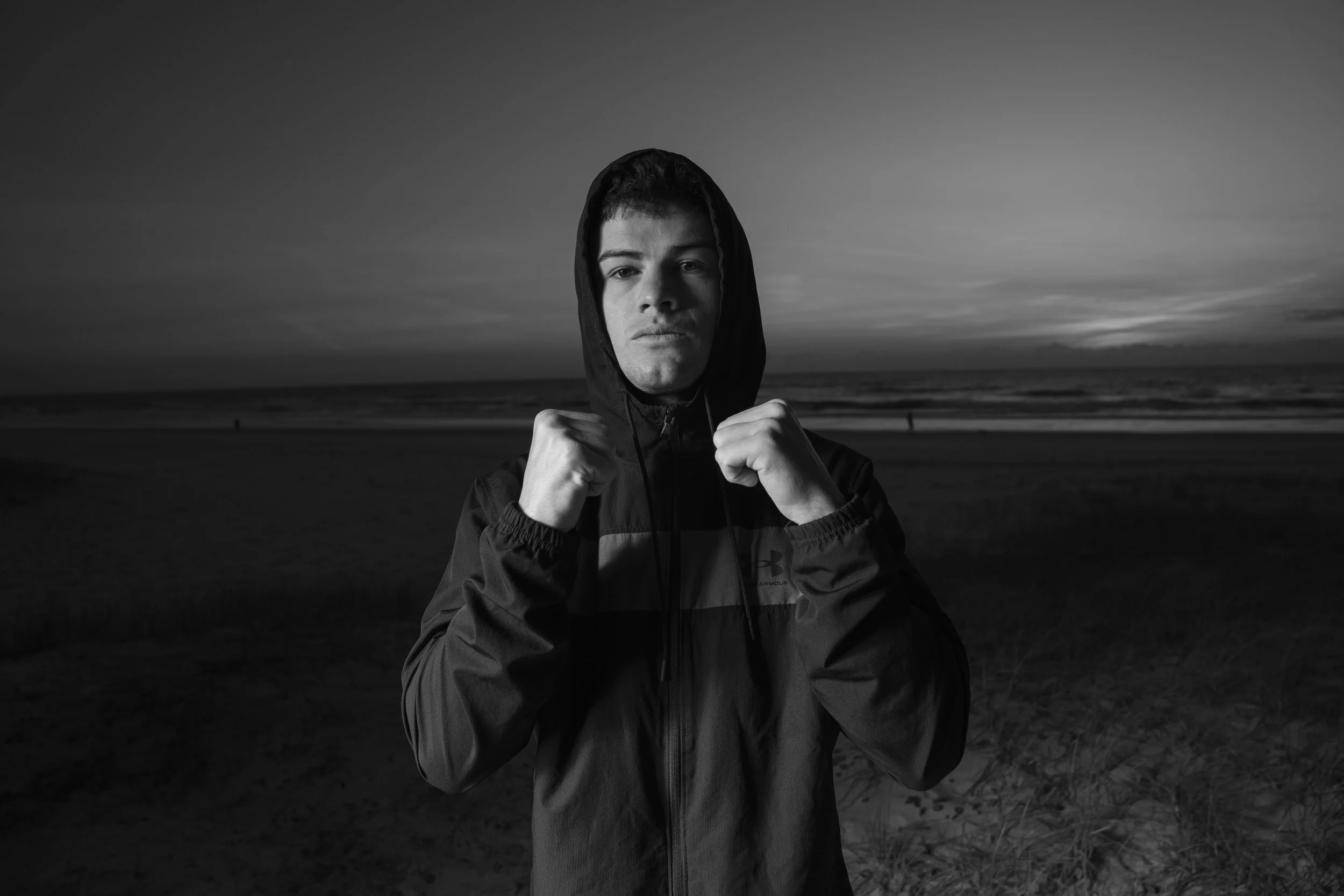 Black and white photo of a man in a hoodie with a boxing pose on a beach at dusk.