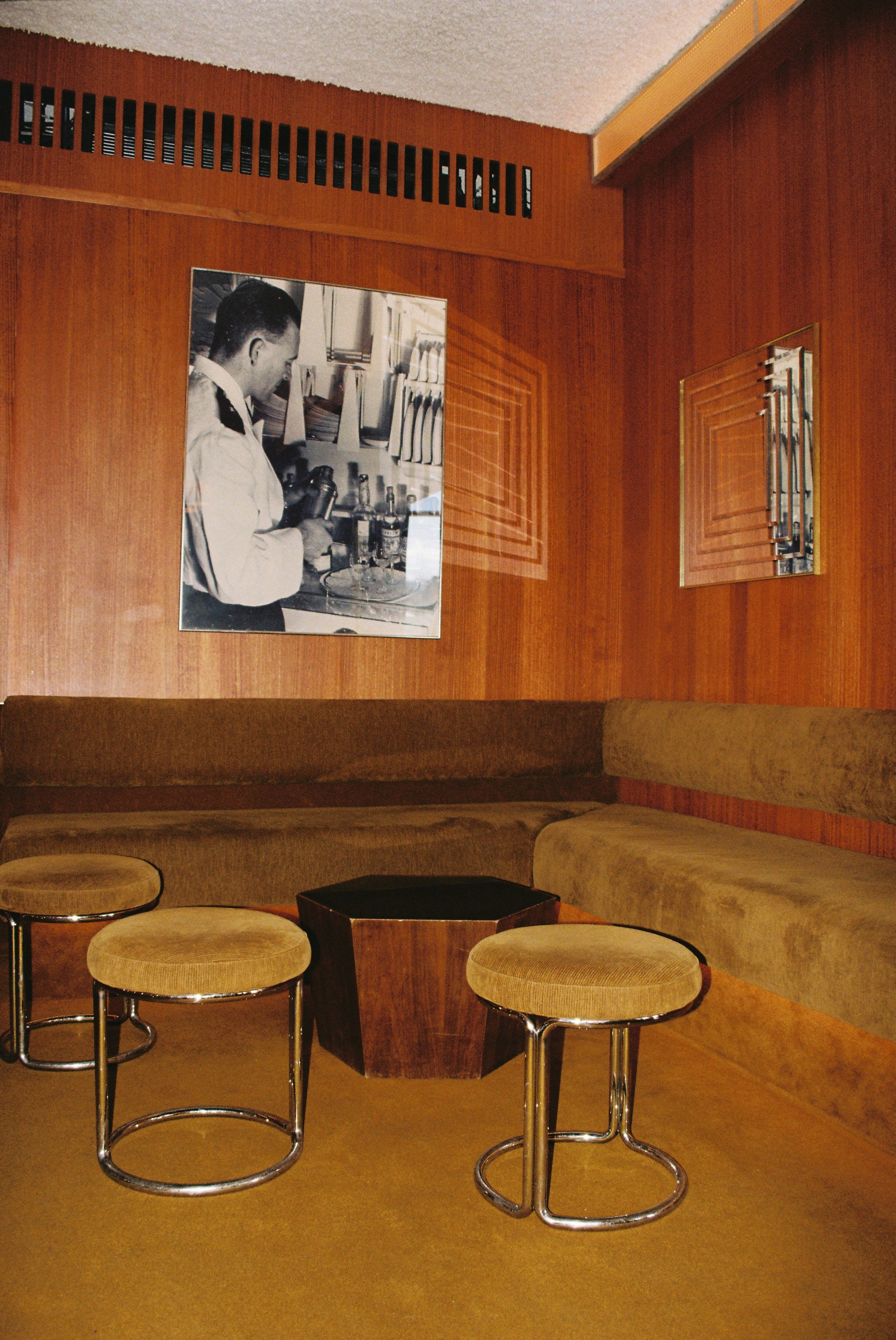 A vintage hotel lobby or lounge with a wood-paneled wall, a black and white photograph of a man using a film camera, and a curved beige sofa. There are round upholstered stools with metal frames, a small black table, and a wall vent.