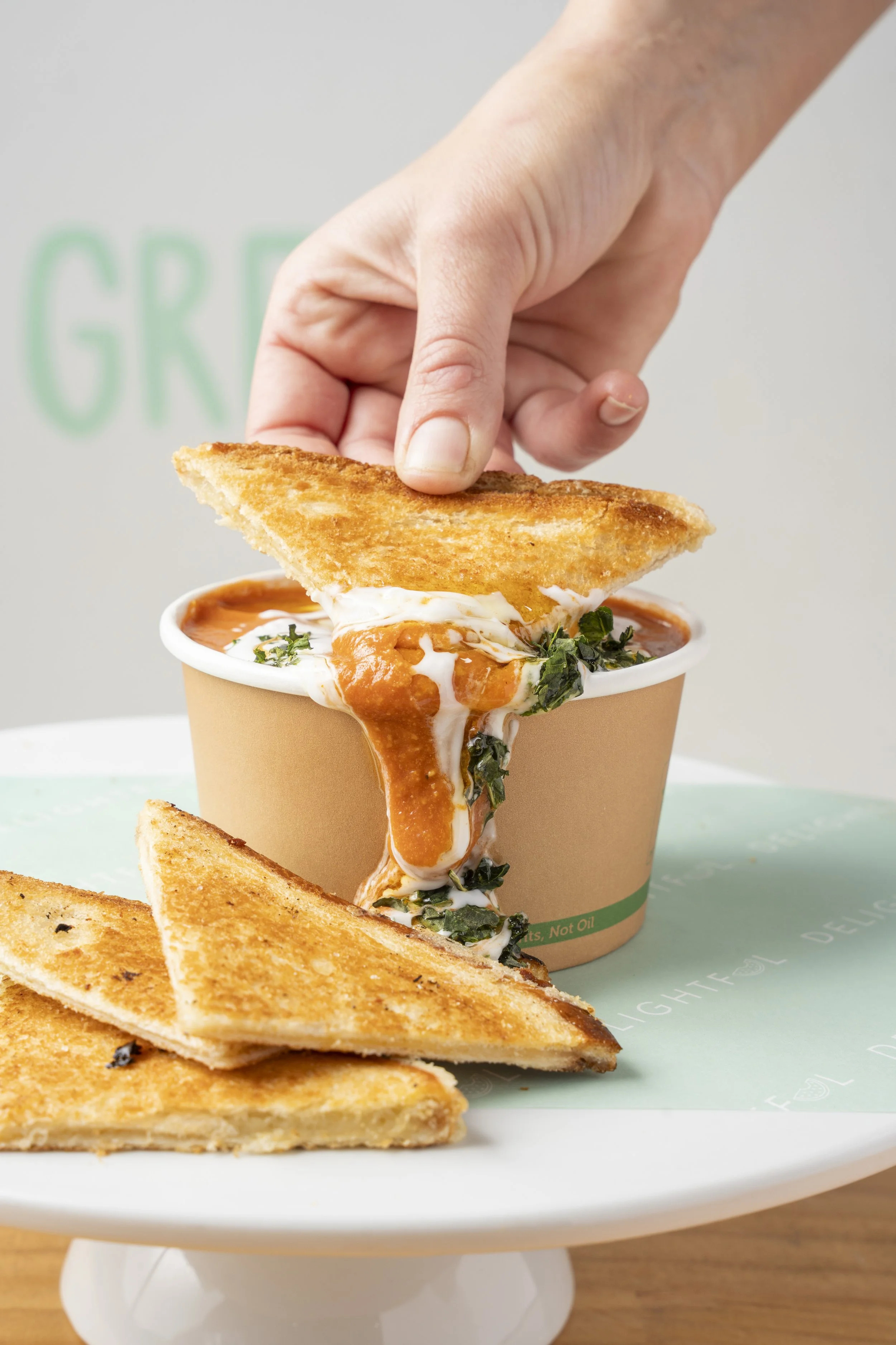 A hand dipping a triangular grilled cheese sandwich with melted cheese, tomato soup, and greens into a paper cup of tomato soup on a white plate. Gold coast food photography by Joey Bailey