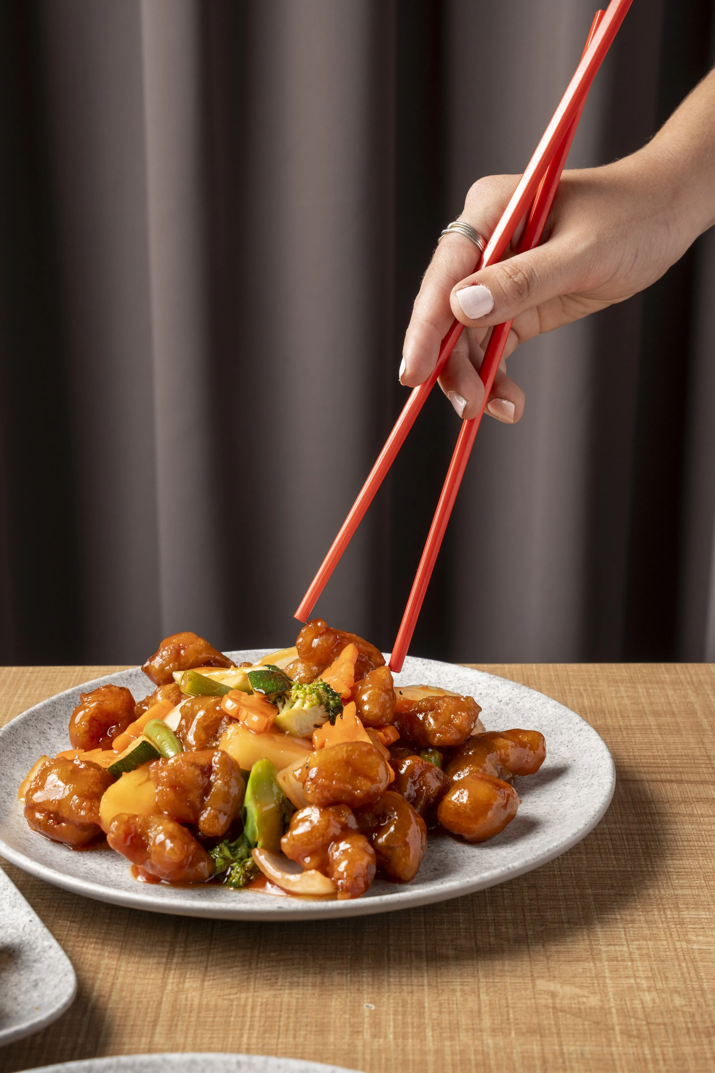 Person using red chopsticks to pick up sweet and sour chicken with vegetables on a white plate. Gold Coast Food Photography by Joey Bailey