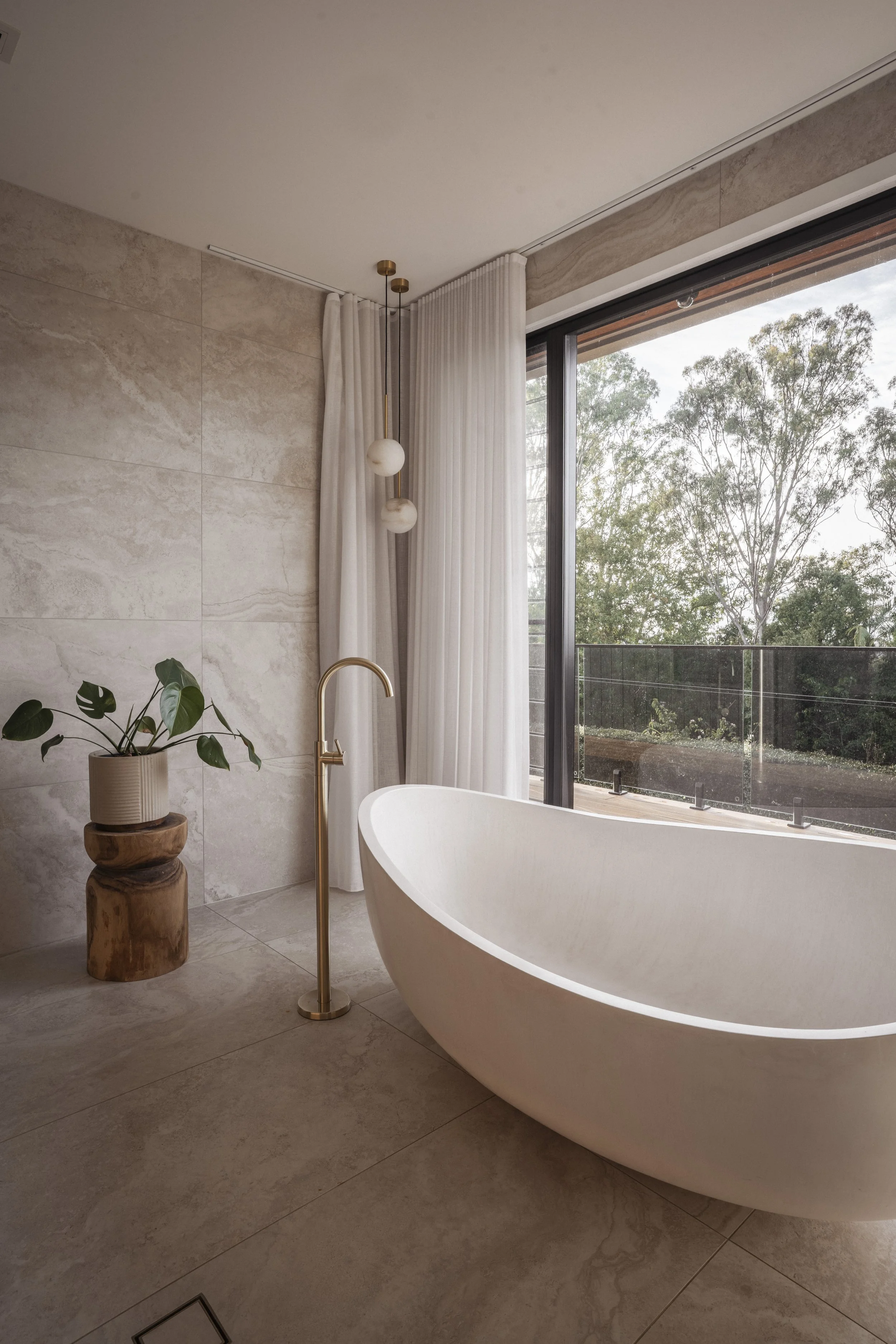 Modern bathroom with a freestanding bathtub near large window, a potted plant on a wooden stand, white curtains, and a minimalist pendant light. Architecture photography by Joey Bailey Gold Coast