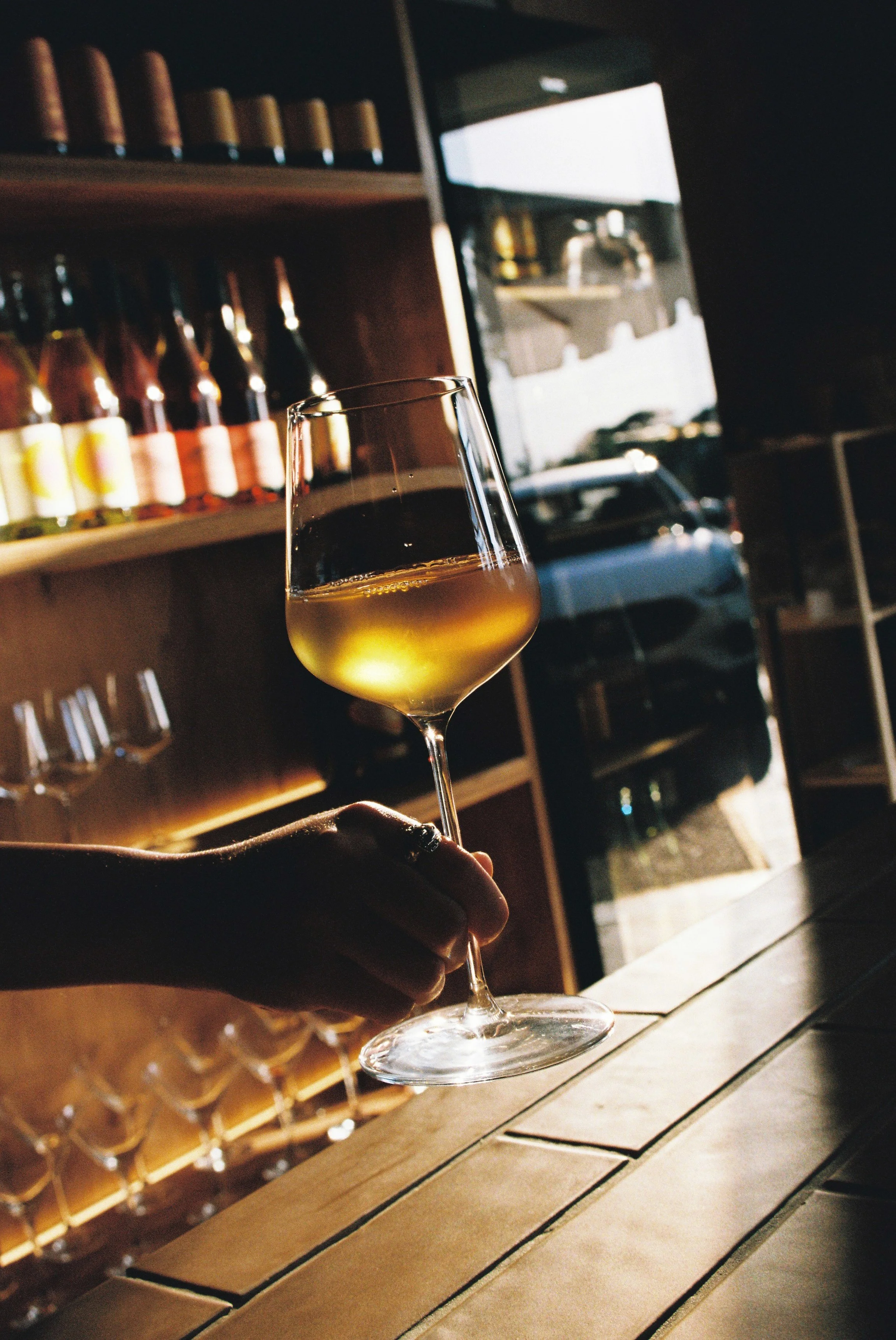 Person holding a glass of white wine in a bar or restaurant with wine bottles on shelves and a large window showing parked cars outside. Gold Coast restaurant food photography 