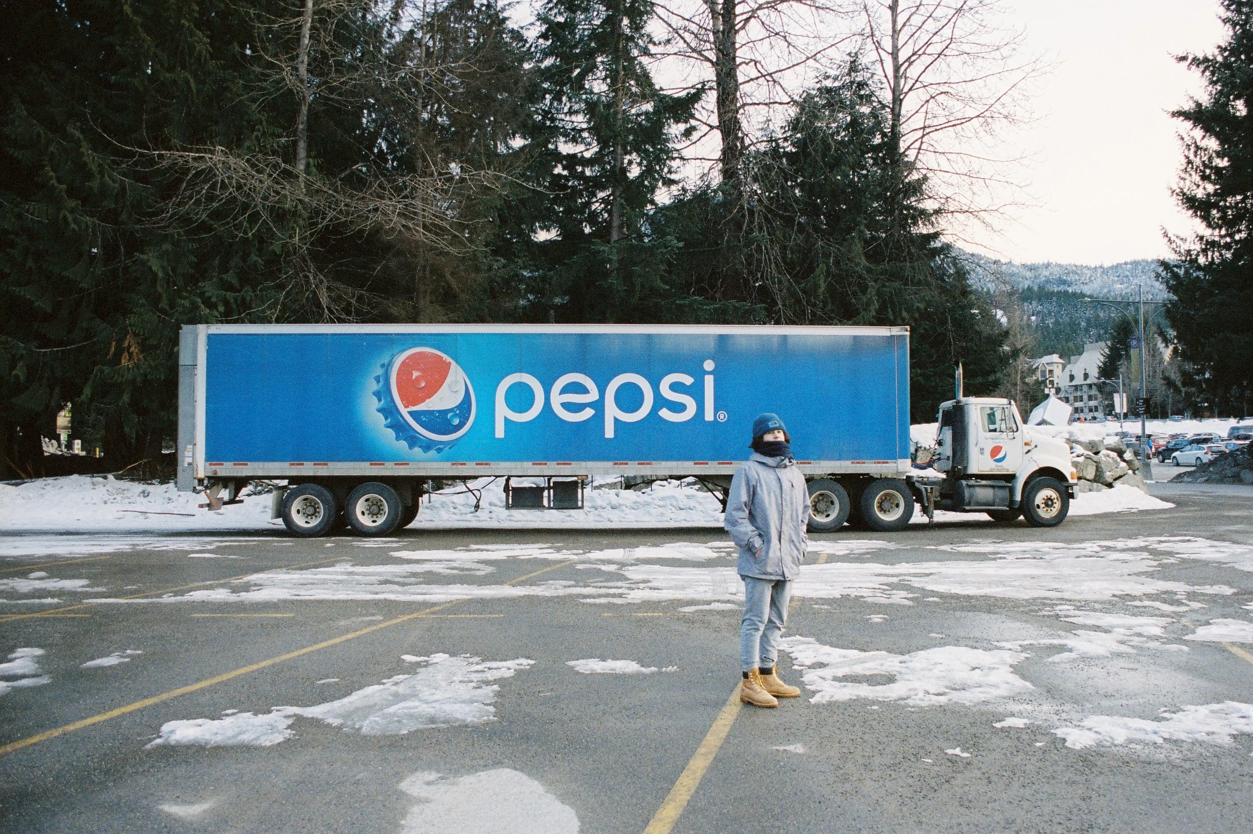 A person in winter clothing stands in front of a Pepsi truck parked in a snowy lot, surrounded by trees.
