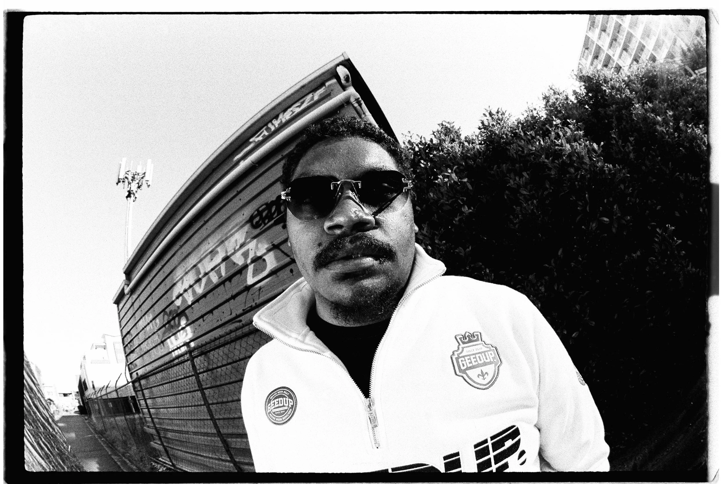 Black and white fisheye photo of a person wearing sunglasses and a jacket with logos, standing next to a graffiti-covered wall and a bush.