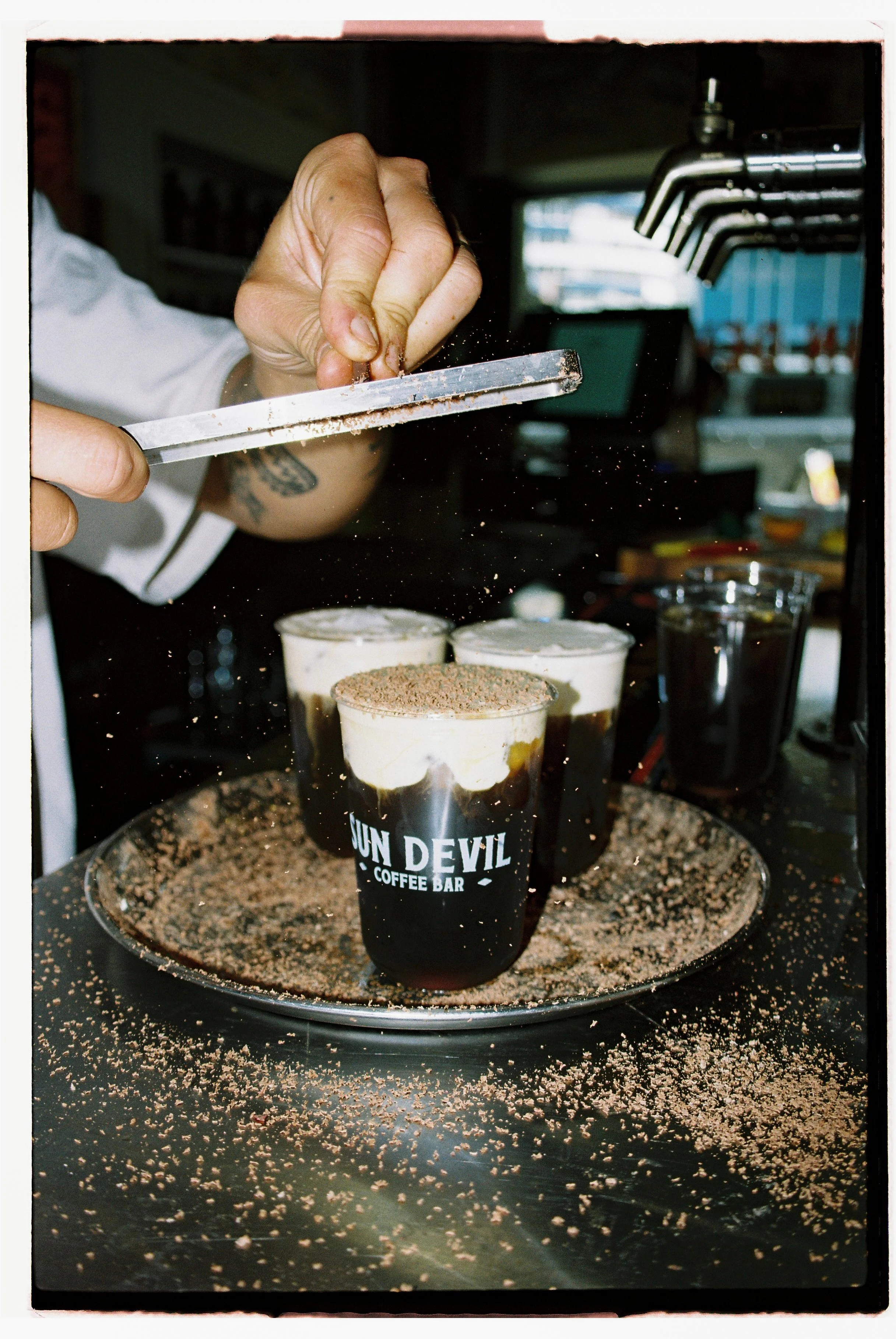 A person is sprinkling a topping onto three cups of iced coffee at a coffee shop counter, with crumbs scattered on the surface and a tray. Sun Devil Cafe NSW by Joey Bailey