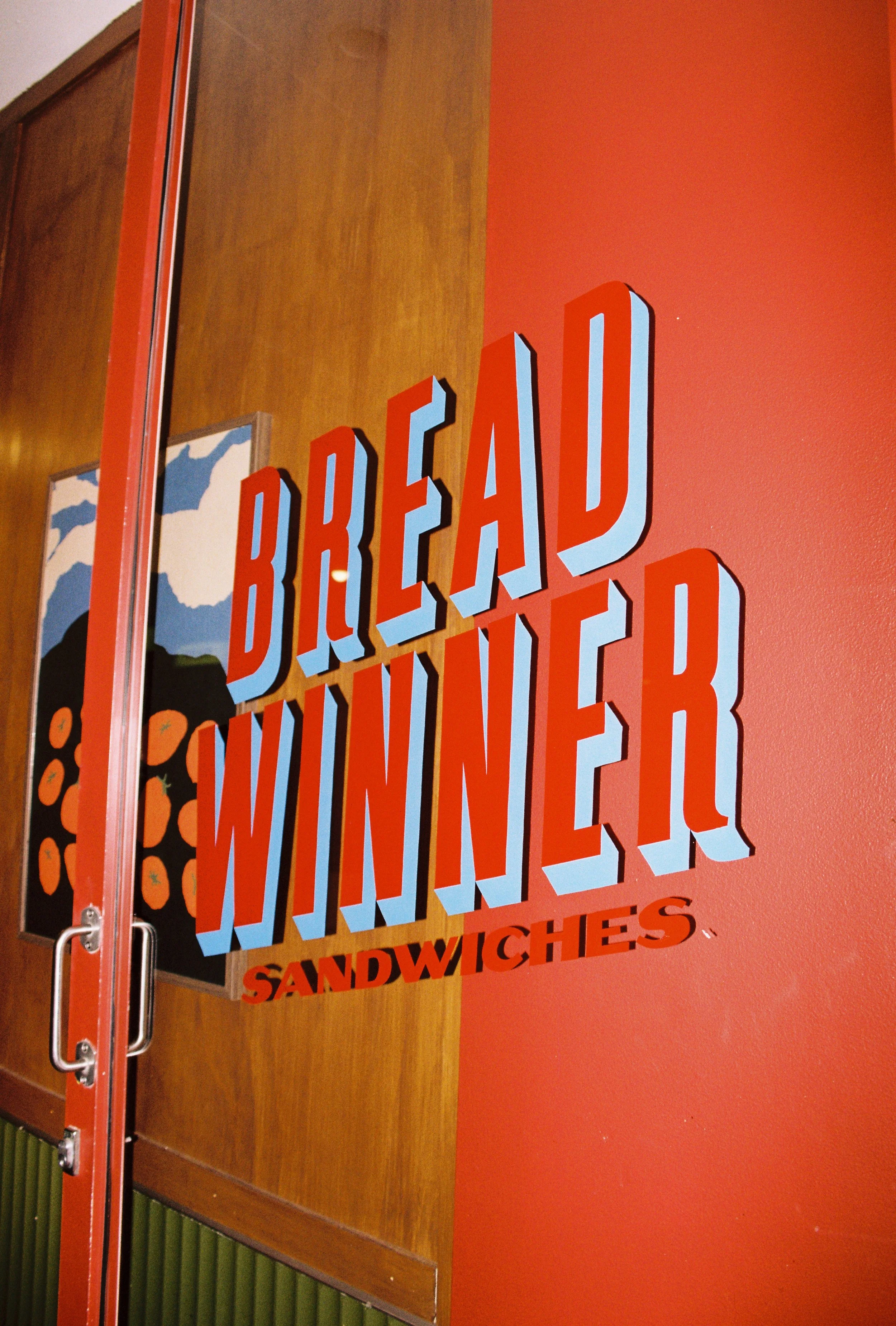 Sign reading 'Break Dau Winner Sandwiches' on a red wall, with a glass door to the left and a wooden panel and artwork in the background. Byron Bay food photography