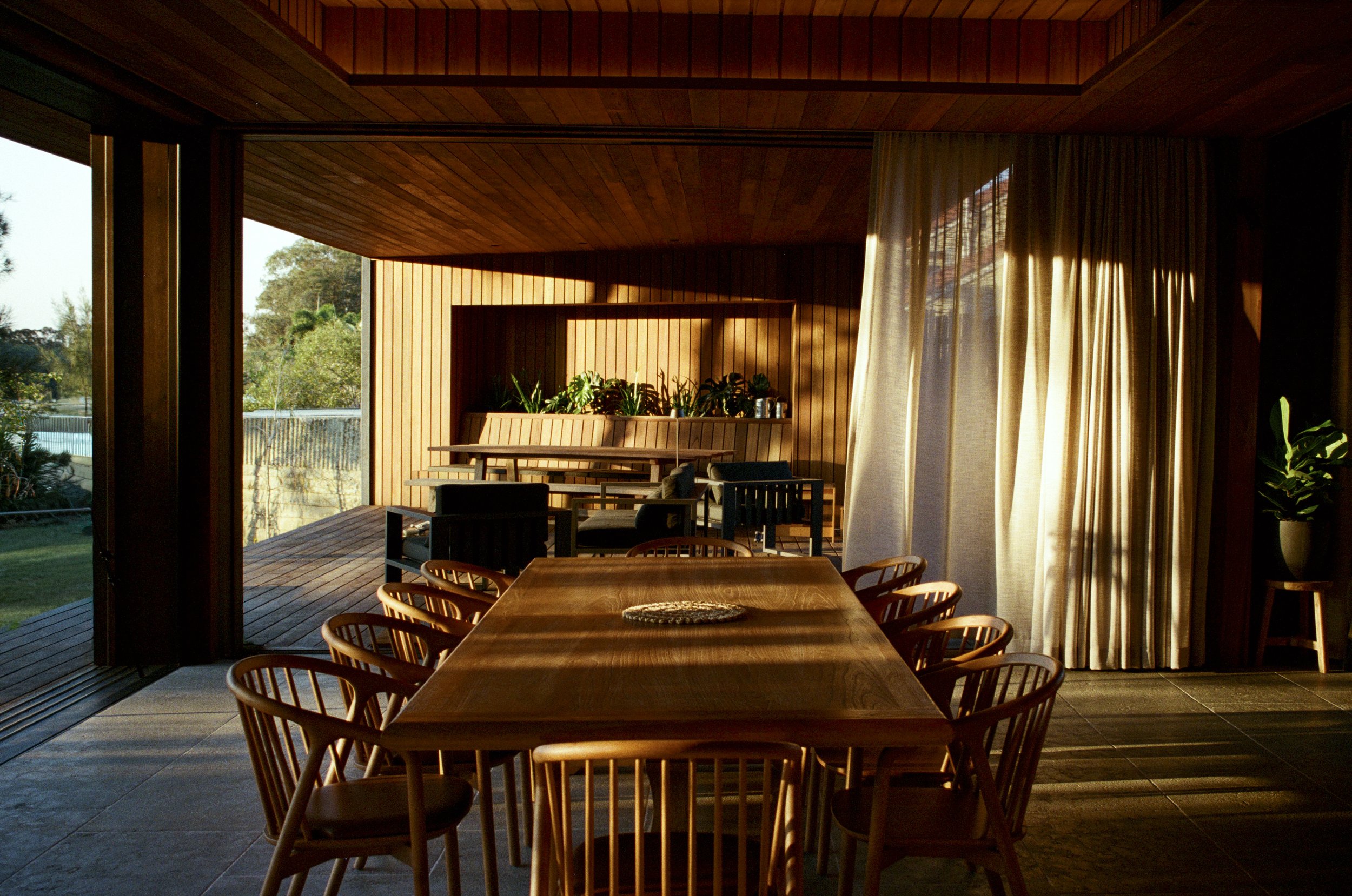 A wooden dining table surrounded by chairs in a sunlit room with large sliding glass doors revealing a deck and backyard. Byron Bay interior photography