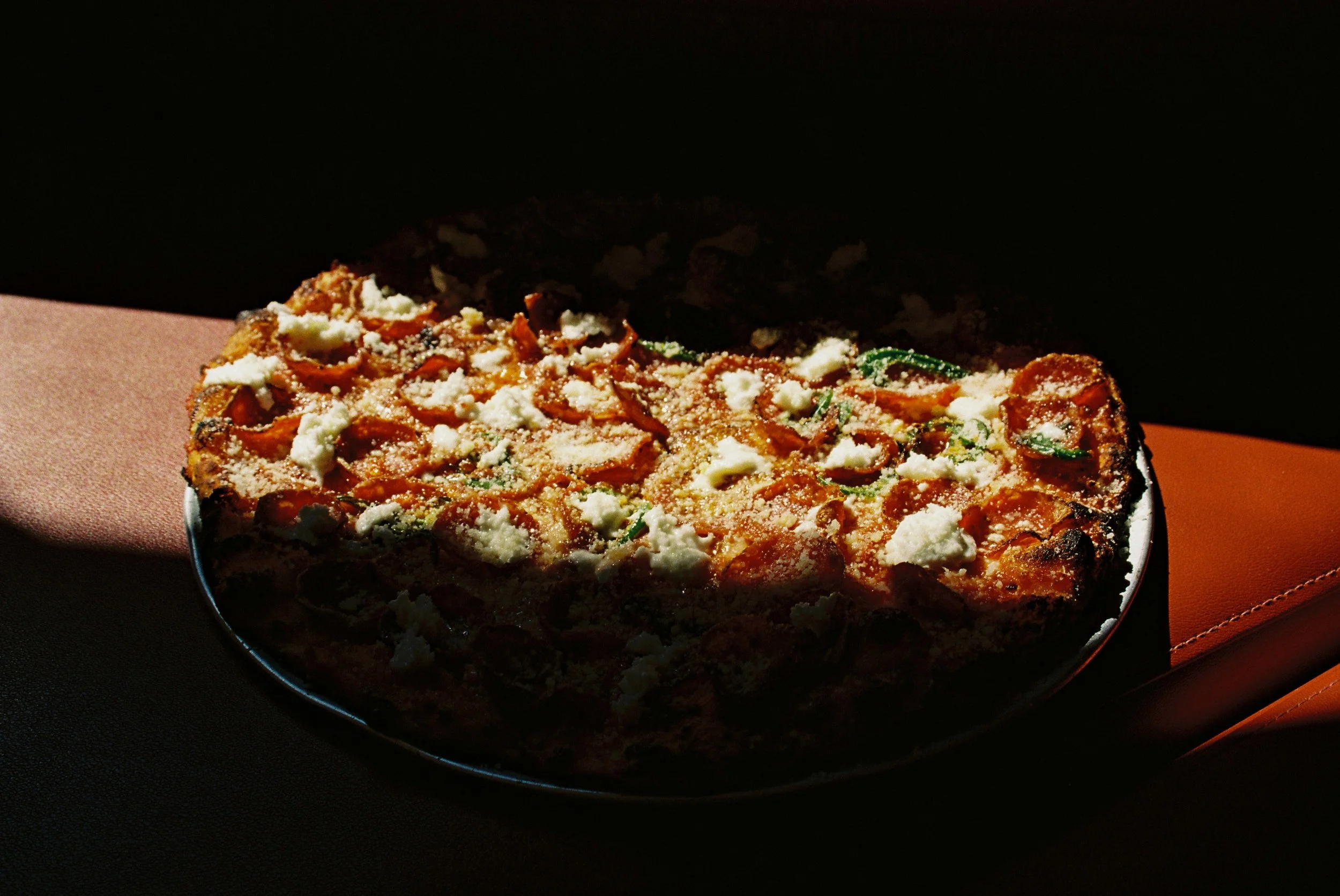 A pizza with toppings including cheese, tomato sauce, and herbs on a round baking pan, placed on a black surface with a pink and orange-colored cloth nearby.  Club Rose Bay Photography
