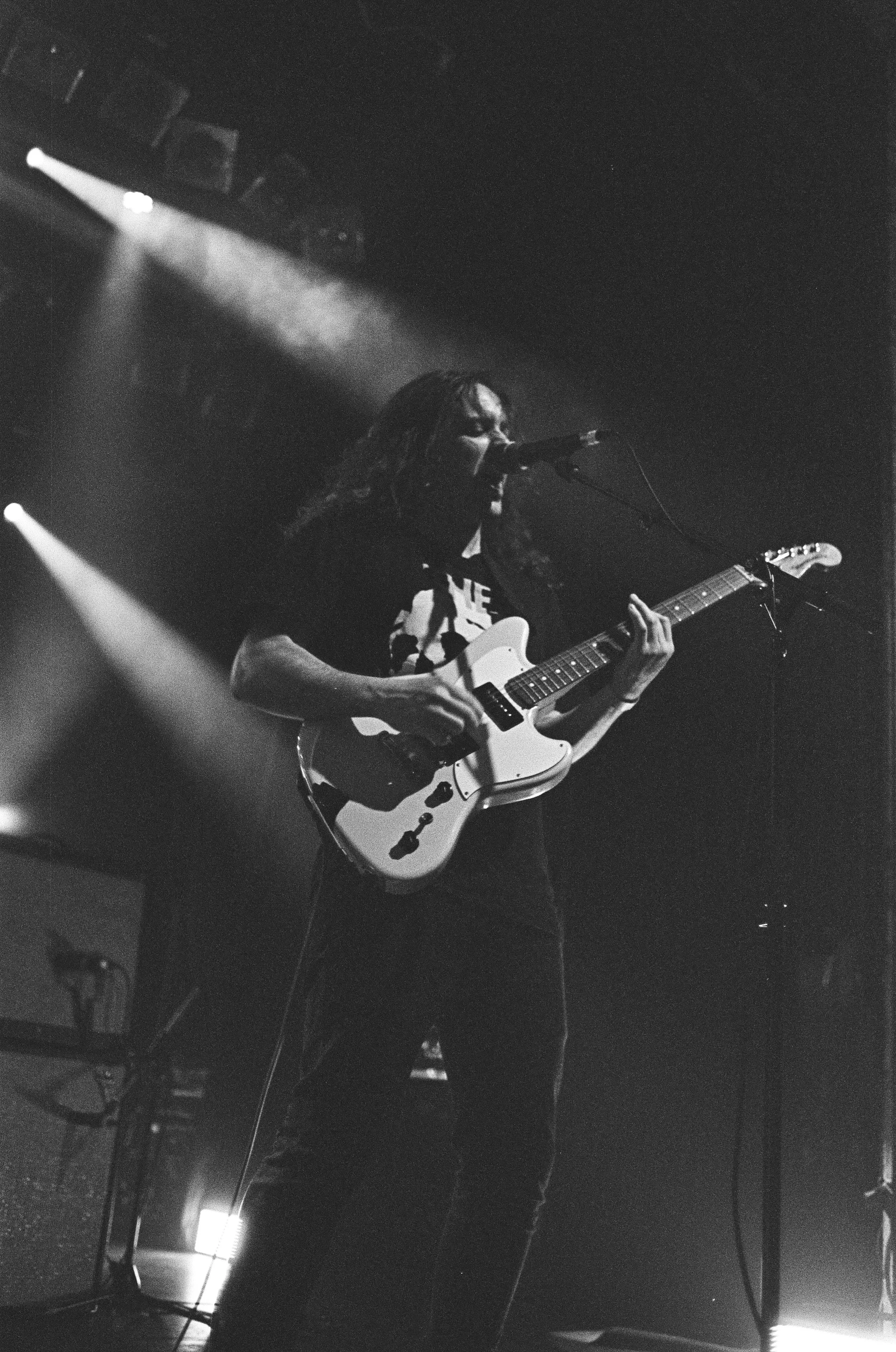 Musician playing electric guitar on stage in black and white, with stage lights in the background.