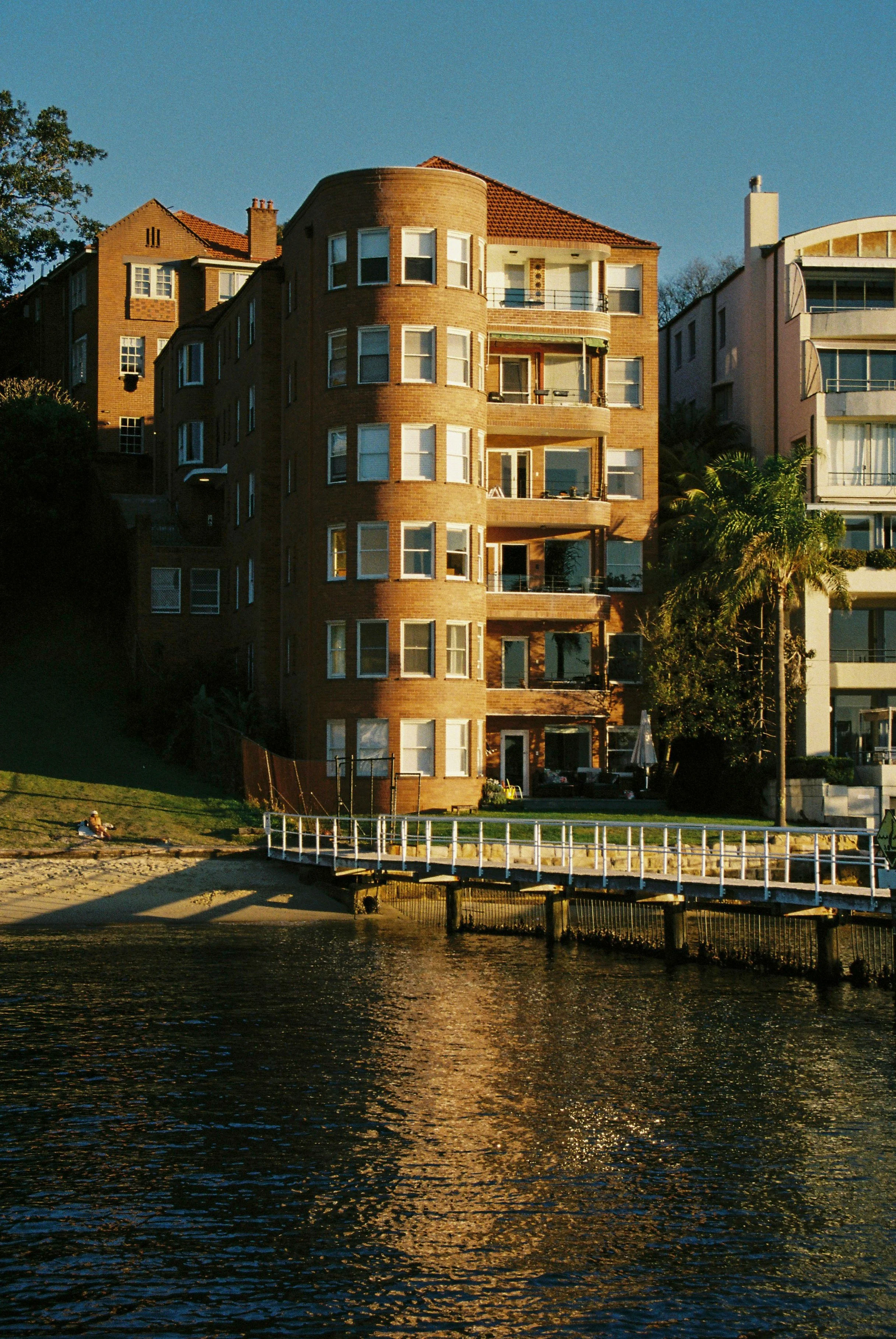 A sunset view of a brick apartment building beside a body of water with a small pier and a person sitting on the grassy area near the water. Garron Tower Art Deco architecture