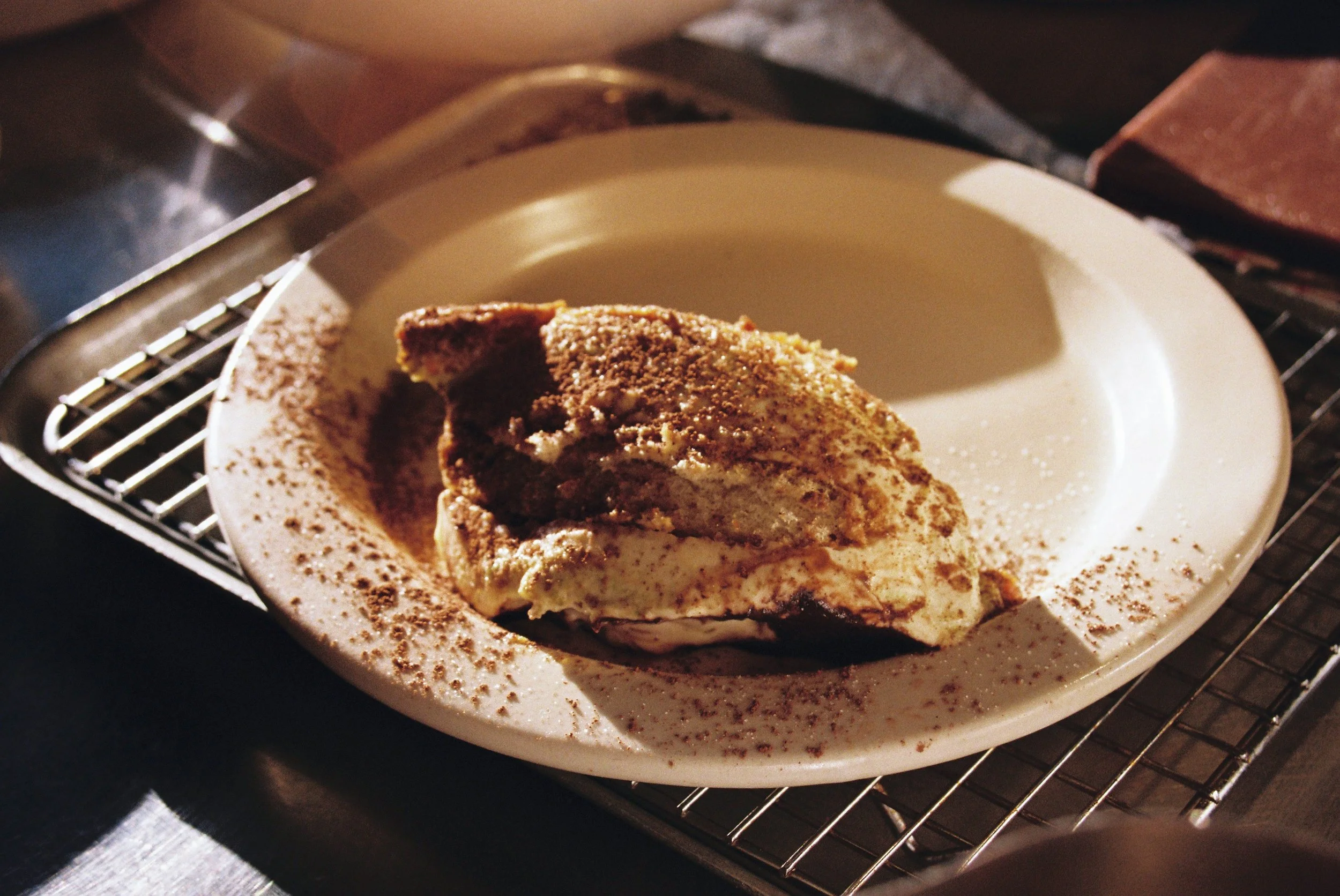 A slice of tiramisu dessert on a white plate, sprinkled with cocoa powder, placed on a cooling rack. Gold Coast Food Photography by Joey Bailey