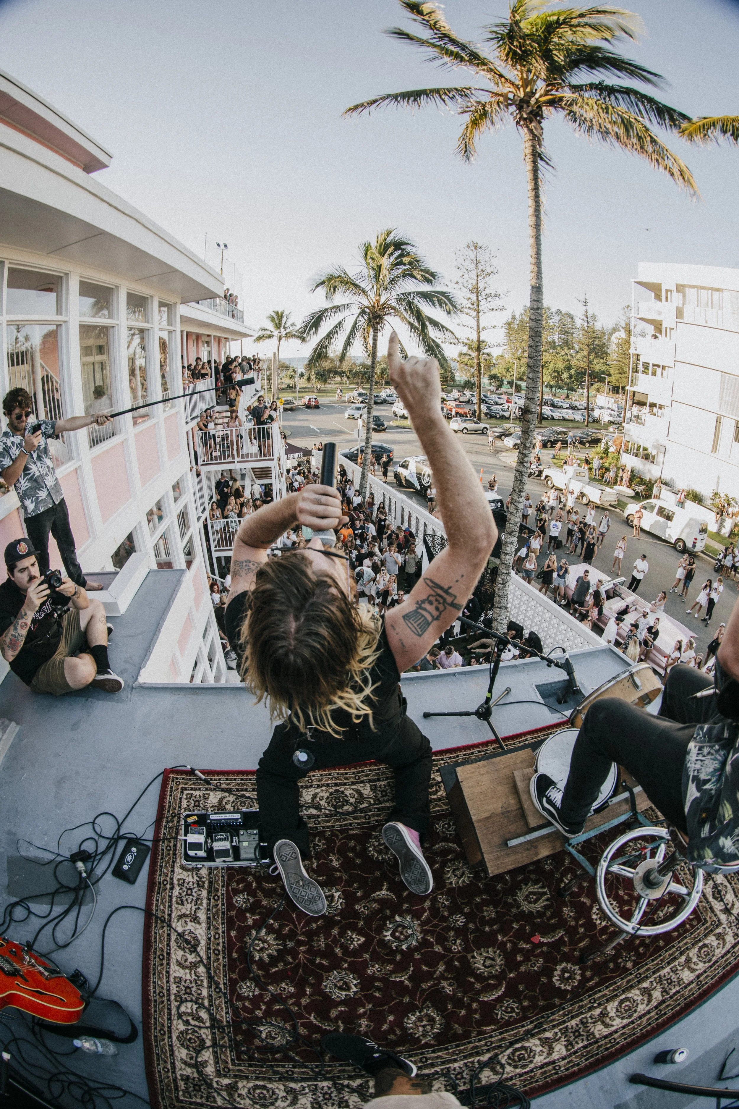 A musician performing on a rooftop with a crowd of spectators below, surrounded by palm trees and a pink building. Several people are capturing the event with cameras.
