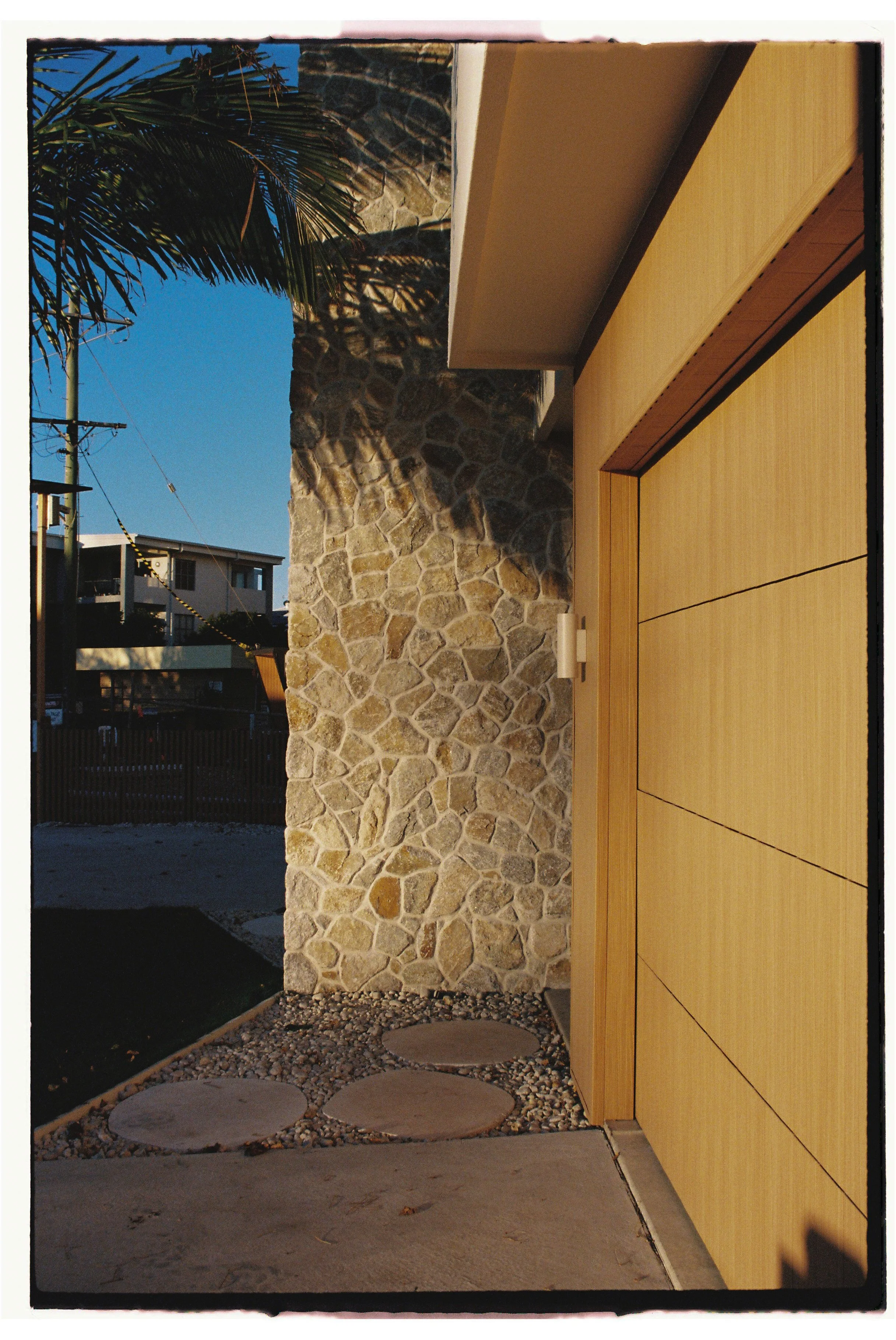 Close-up of modern house exterior showing a stone wall, wooden sliding door, and entry pathway with large circular stepping stones and small gravel. Architecture photography Gold Coast by Joey Bailey