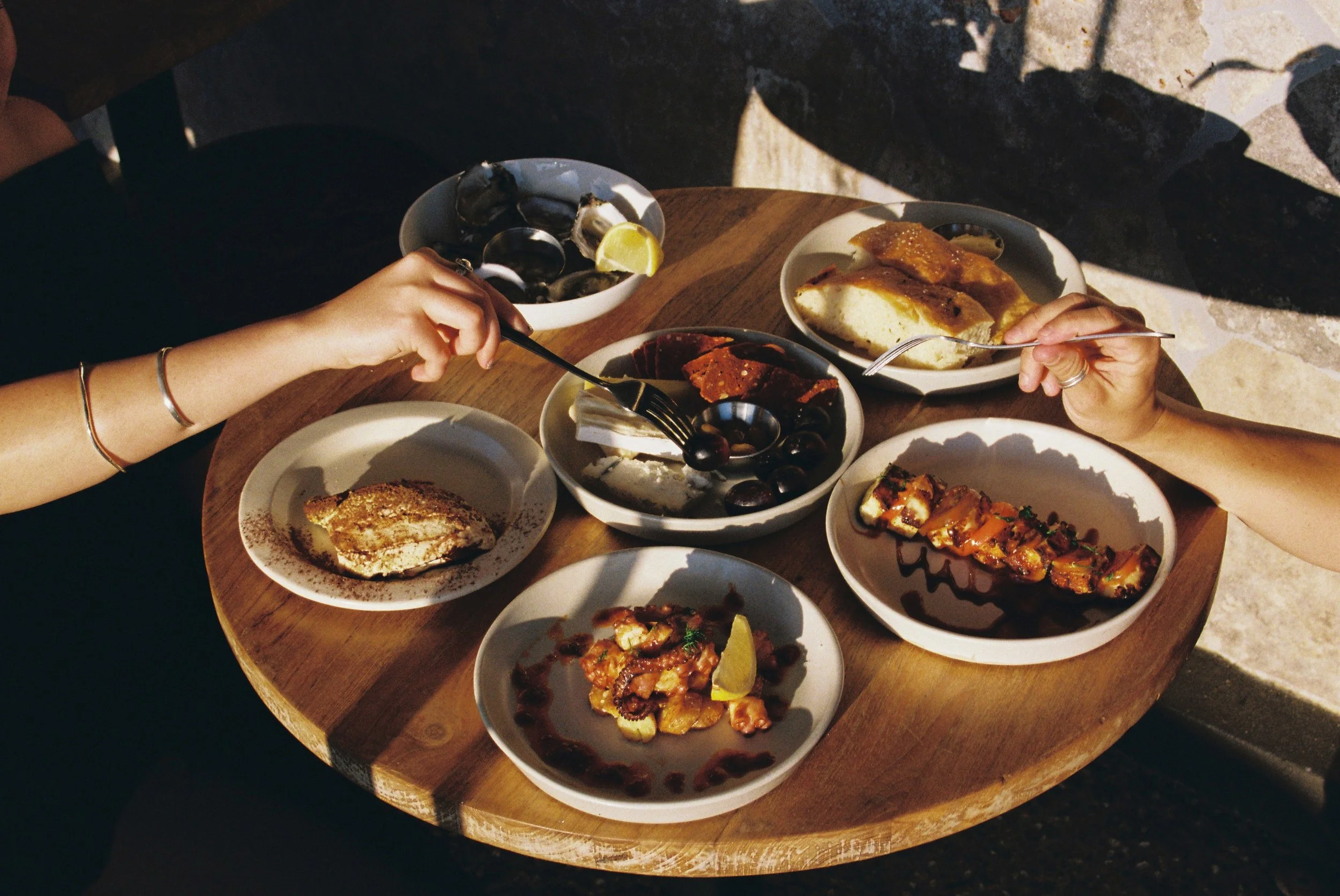 Two people sharing a round wooden table with various plates of food, moustache mermaid beach photography