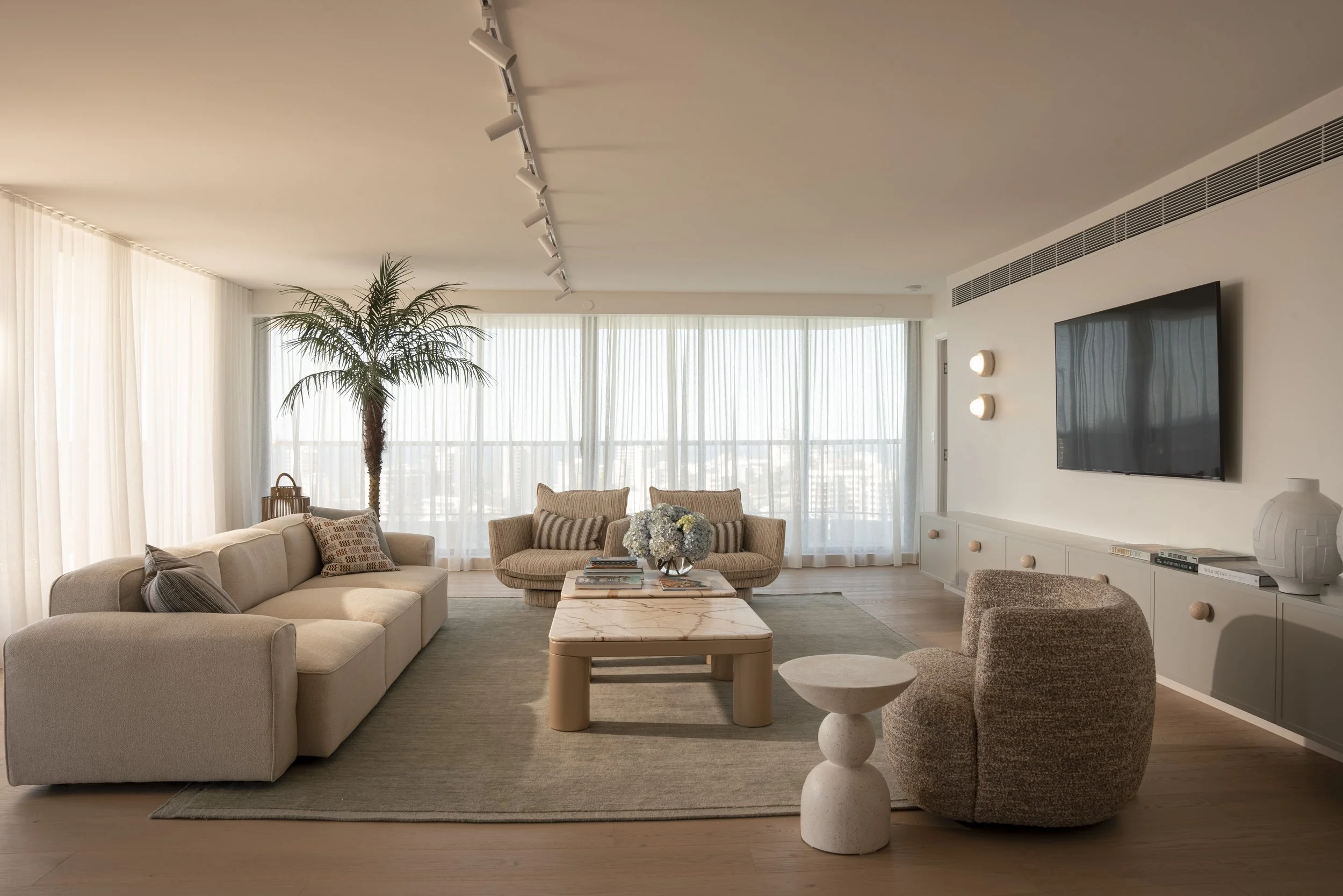 A spacious living room with beige and neutral-toned furniture, including a sofa, armchairs, and a coffee table, with large windows covered by sheer curtains, Gold Coast Architecture photography by Joey Bailey.
