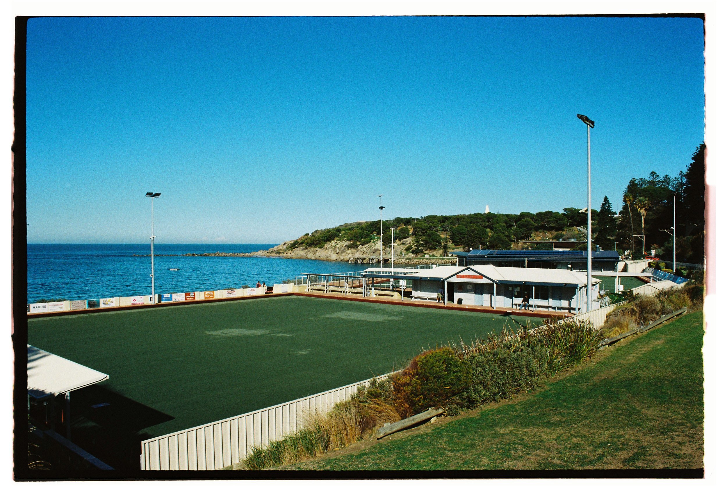A cricket ground with a green pitch by the sea, surrounded by a white fence, with a clubhouse, tall stadium lights, and a coastal landscape with trees and hills in the background.