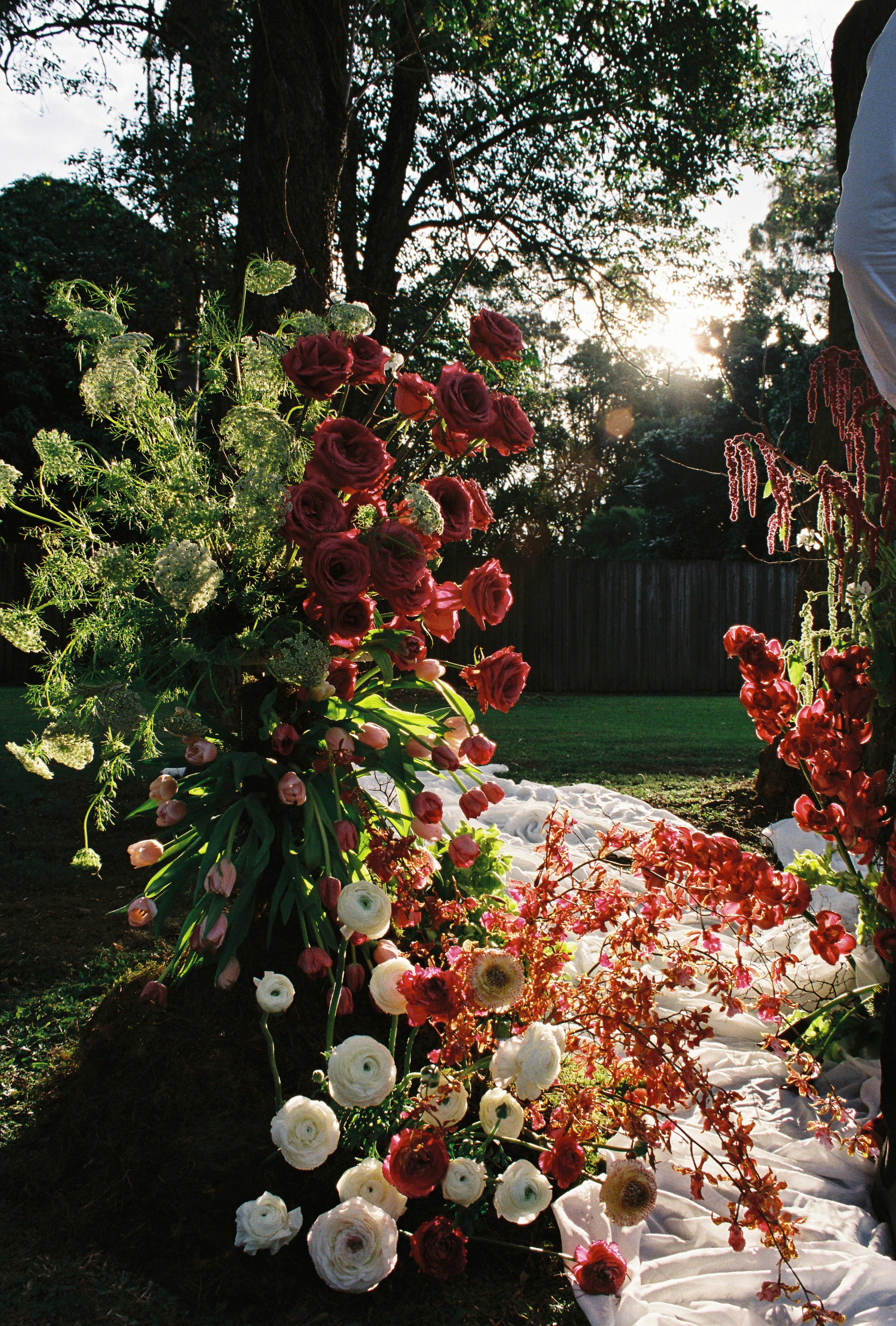 A floral arrangement with pink, white, and red flowers, with white fabric draped on the ground, outdoors in a garden with trees and sunlight in the background.