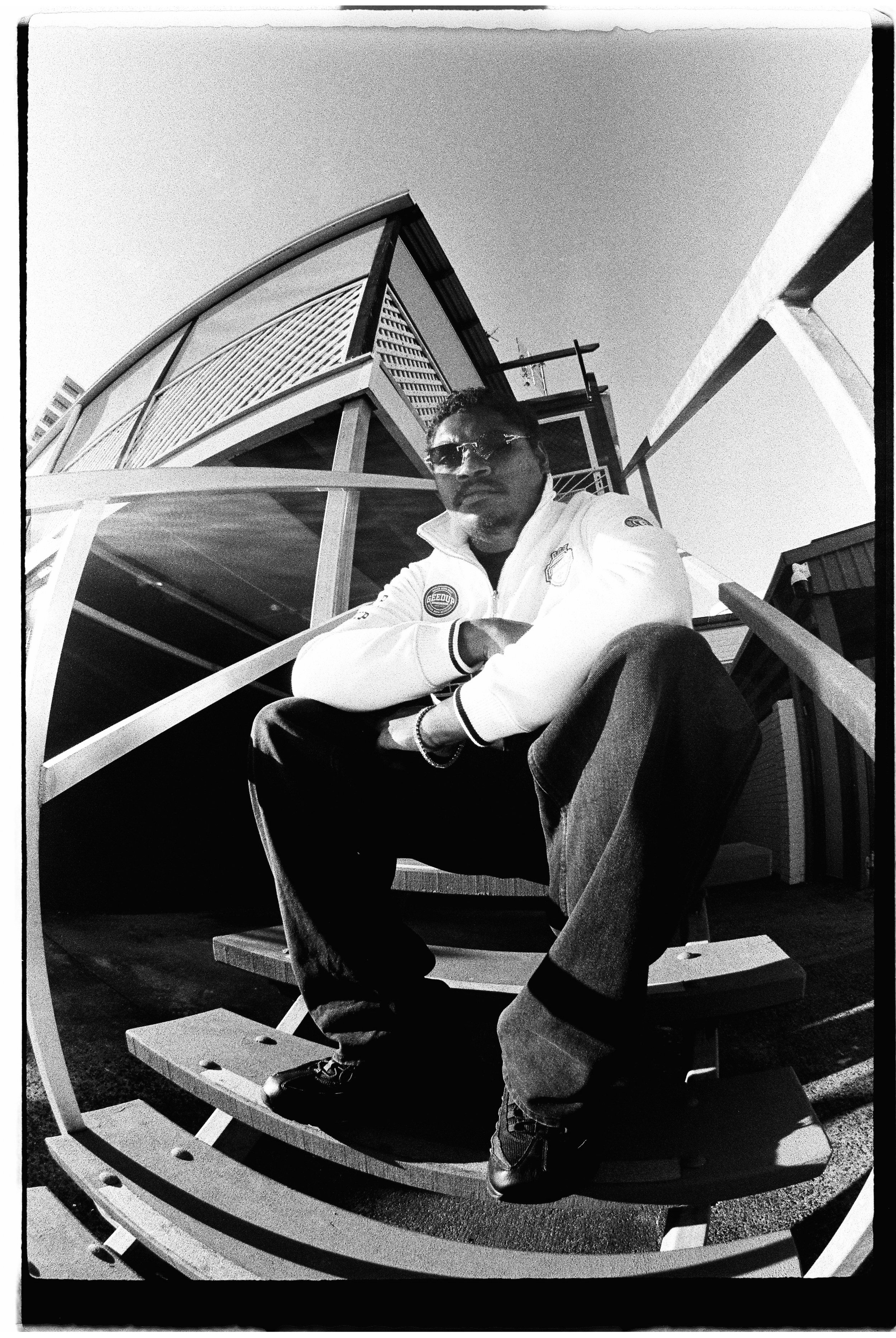 Black and white photo of a person sitting on stairs, wearing a white jacket and sunglasses, with a building in the background.