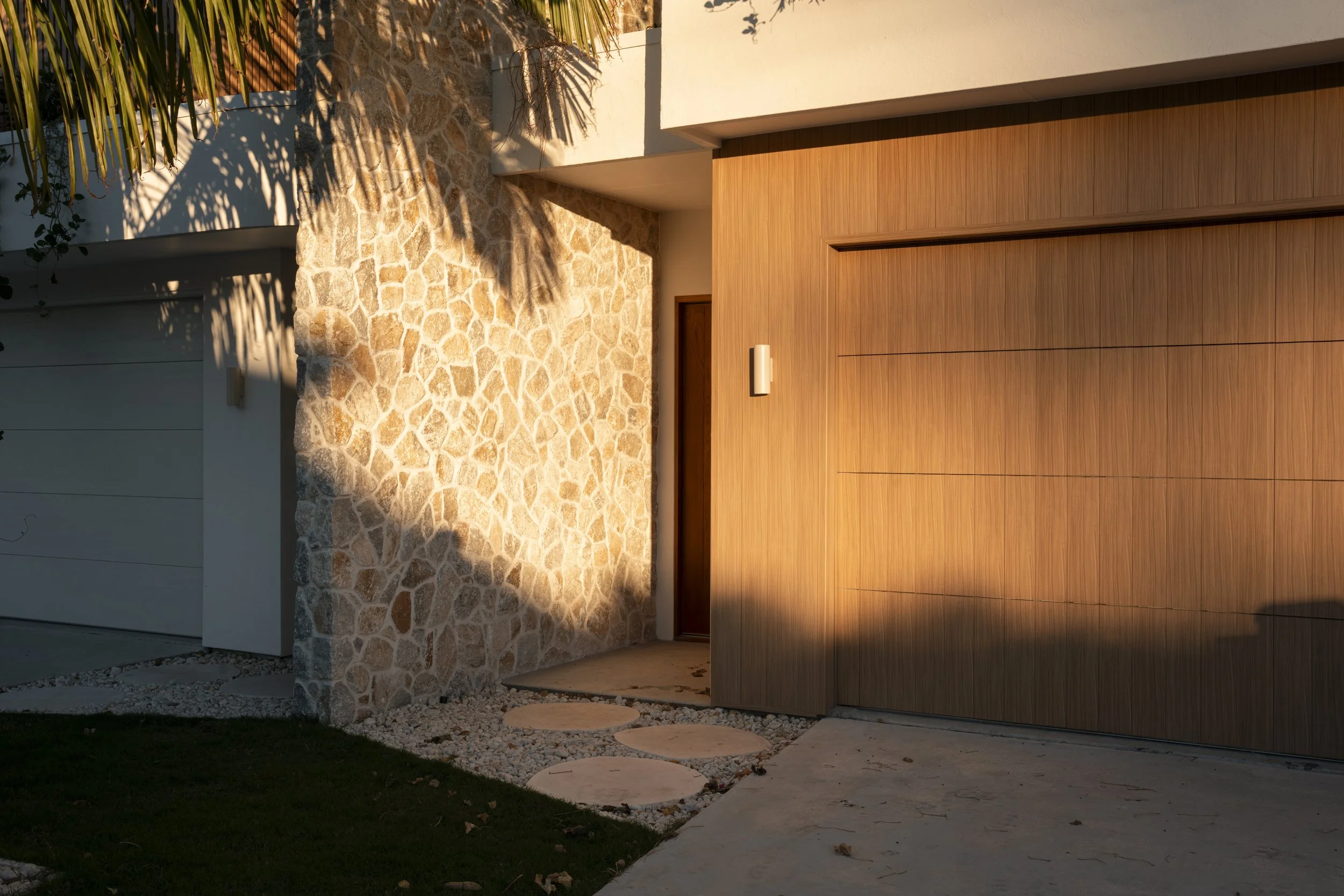 The front entrance of a modern house during sunset, showing a stone wall, wooden garage door, and stepping stones leading to the door. Architectural photography by Joey Bailey, Gold Coast