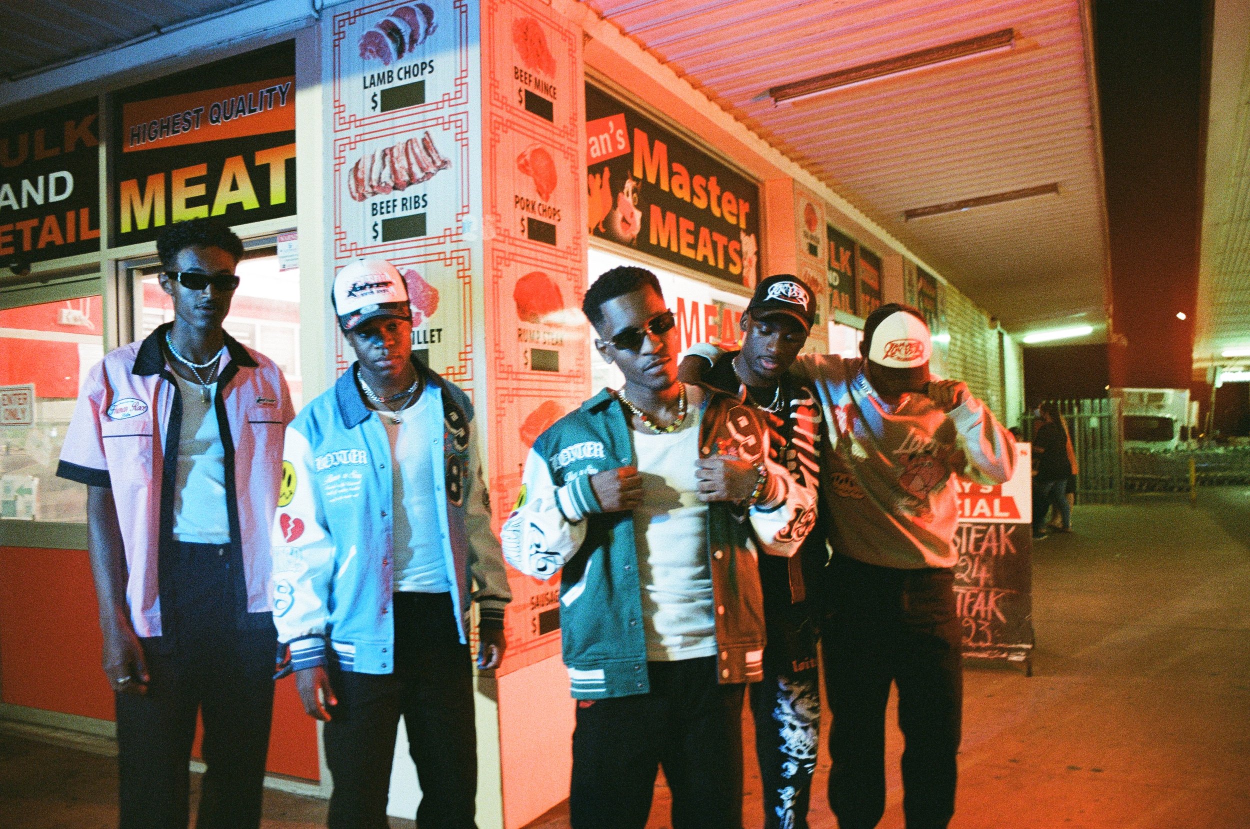 Group of five stylish young men standing in front of a meat market at night, wearing colorful jackets, sunglasses, and caps, with neon and red lighting effects.
