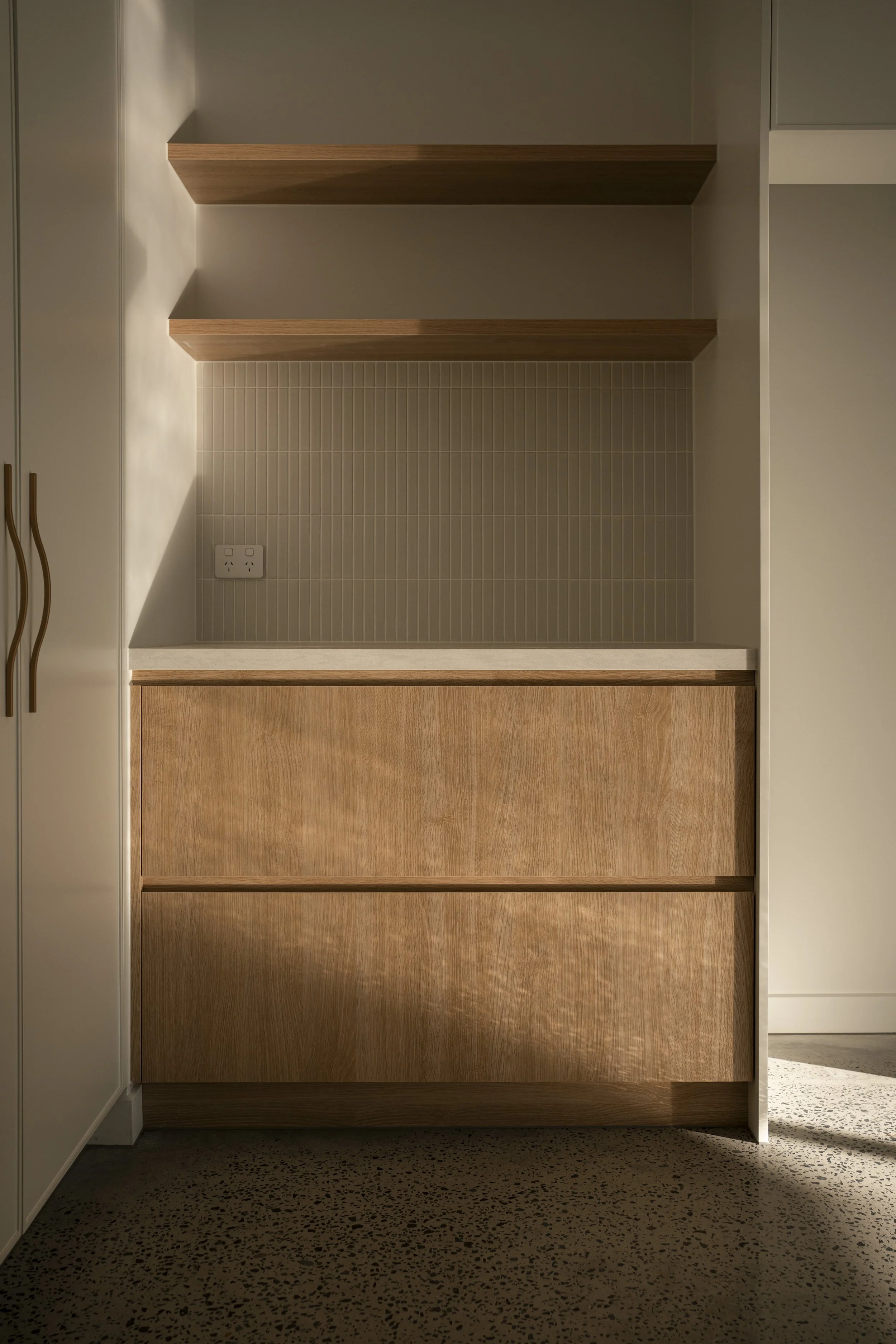 Corner of a room with two hanging wooden shelves, a power outlet, and wooden drawers below a beige tiled wall. Architectural photography by Joey Bailey, Gold Coast