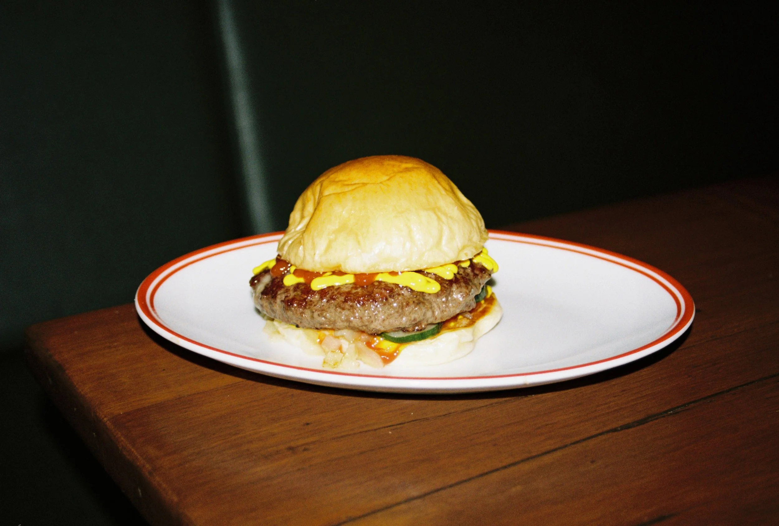 A cheeseburger with a beef patty, cheese, and toppings on a bun, served on a white plate with red trim, placed on a wooden surface. Byron Bay food photography