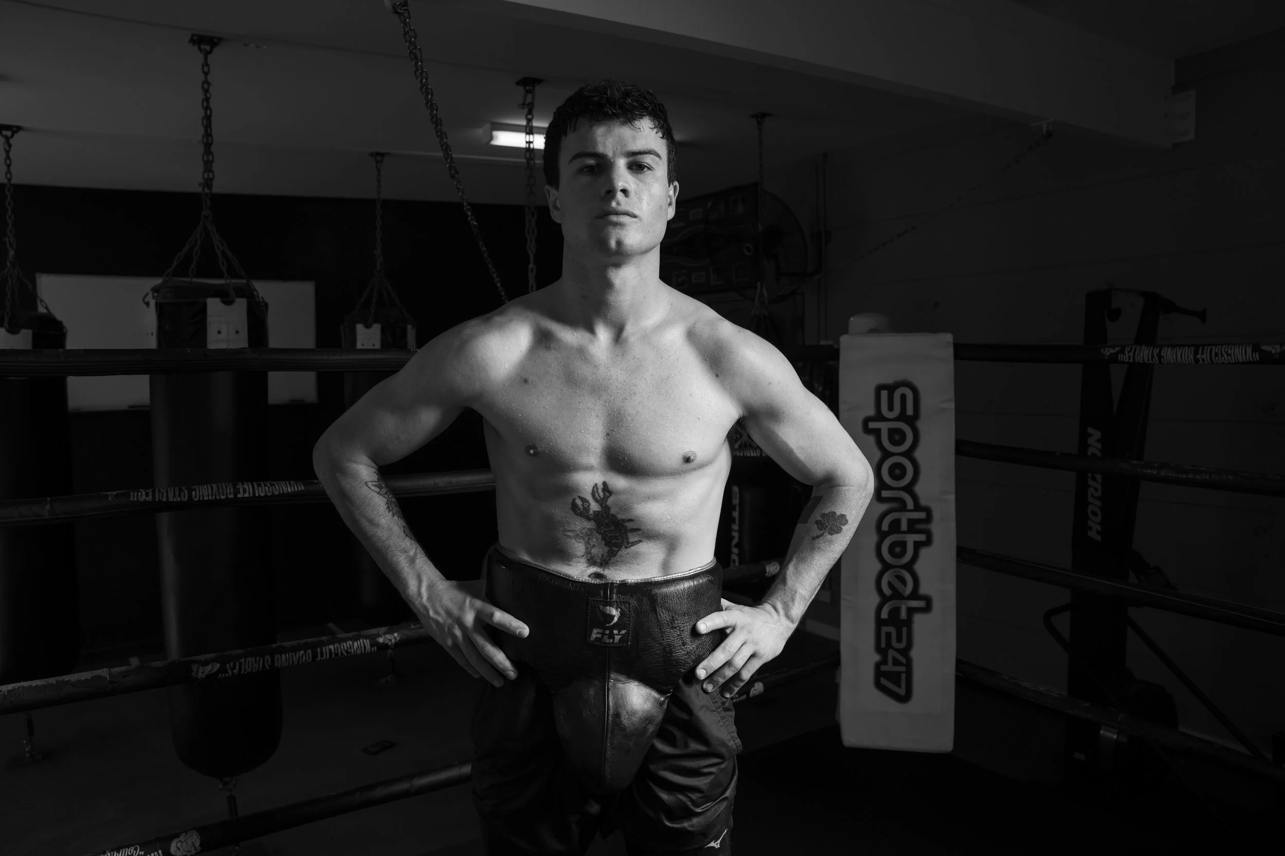 Shirtless boxer posing in a boxing gym, wearing protective gear, with punching bags and ring ropes in the background.