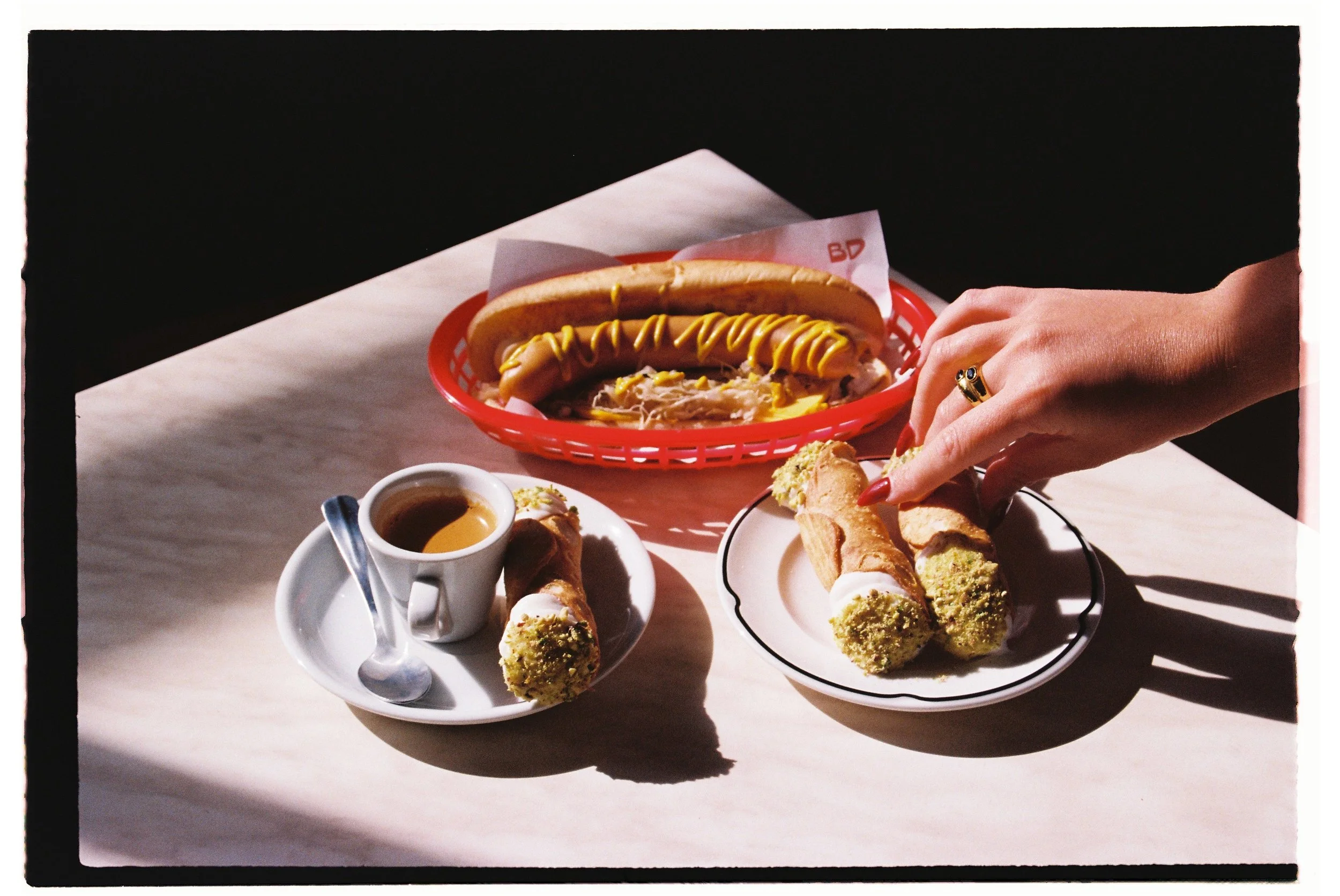 A table with hot dogs, a cup of coffee, and other snacks. A person's hand is reaching for a hot dog on a plate. food photography by Joey Bailey