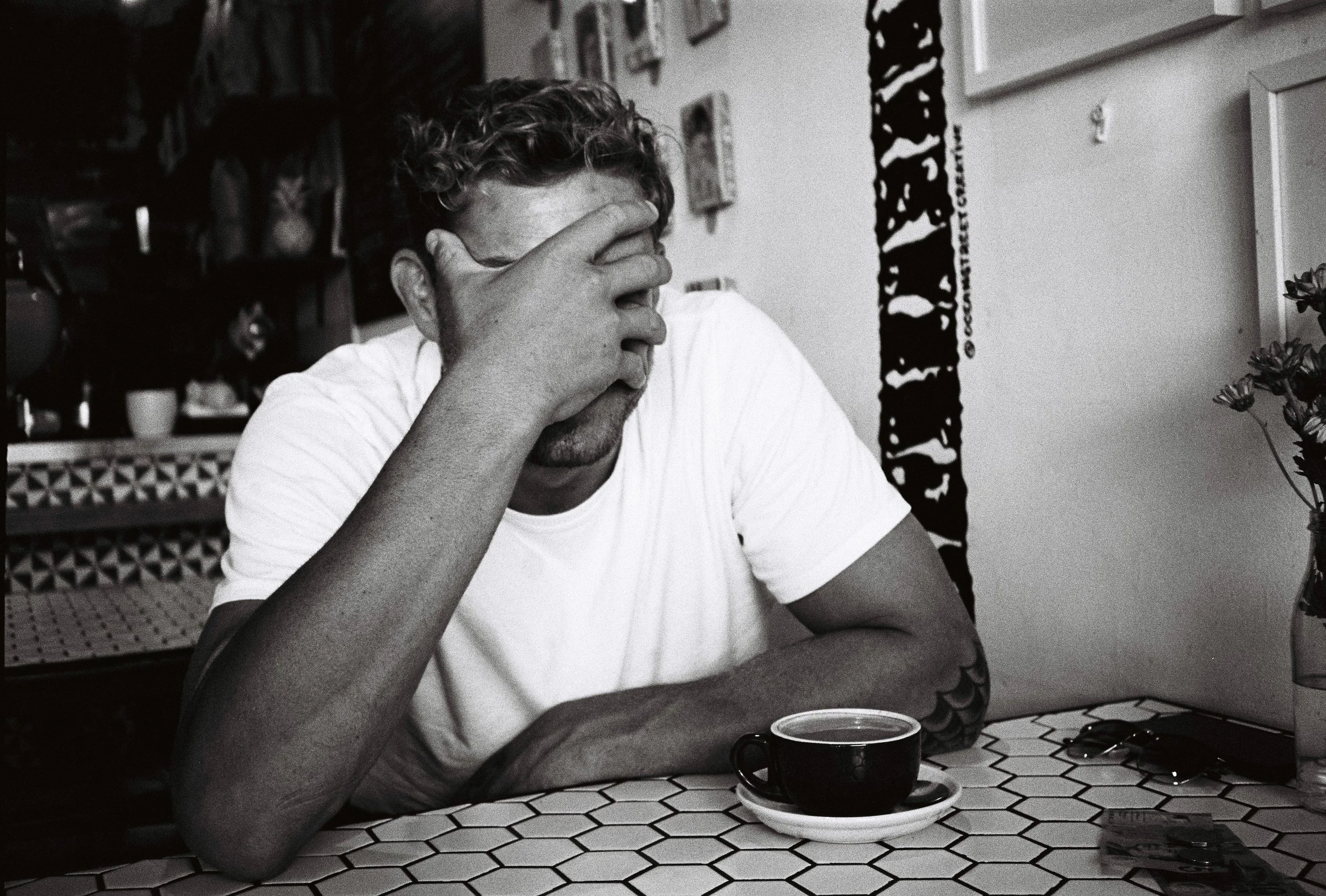 Black and white photo of a person sitting at a table in a cafe, wearing a white t-shirt, with their hand covering their face, and a cup of coffee on the table.