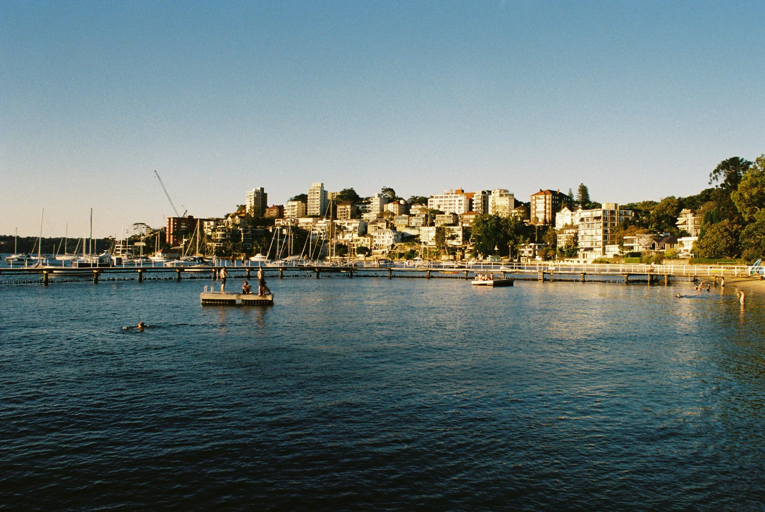 A marina with sailboats docked and a hillside cityscape with modern residential buildings in the background, under a clear blue sky. Double Bay architecture photography