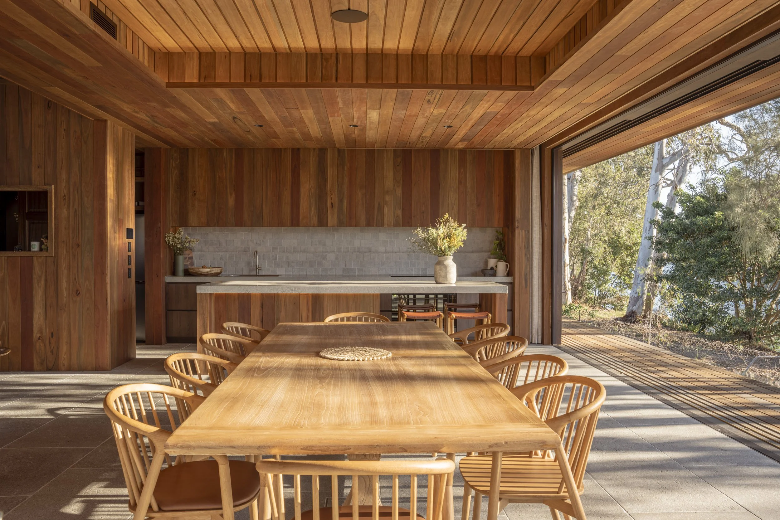 Modern dining area with wooden table and chairs, kitchen with light gray backsplash, and large windows overlooking trees. Currumbin Architecture Photography Joey Bailey