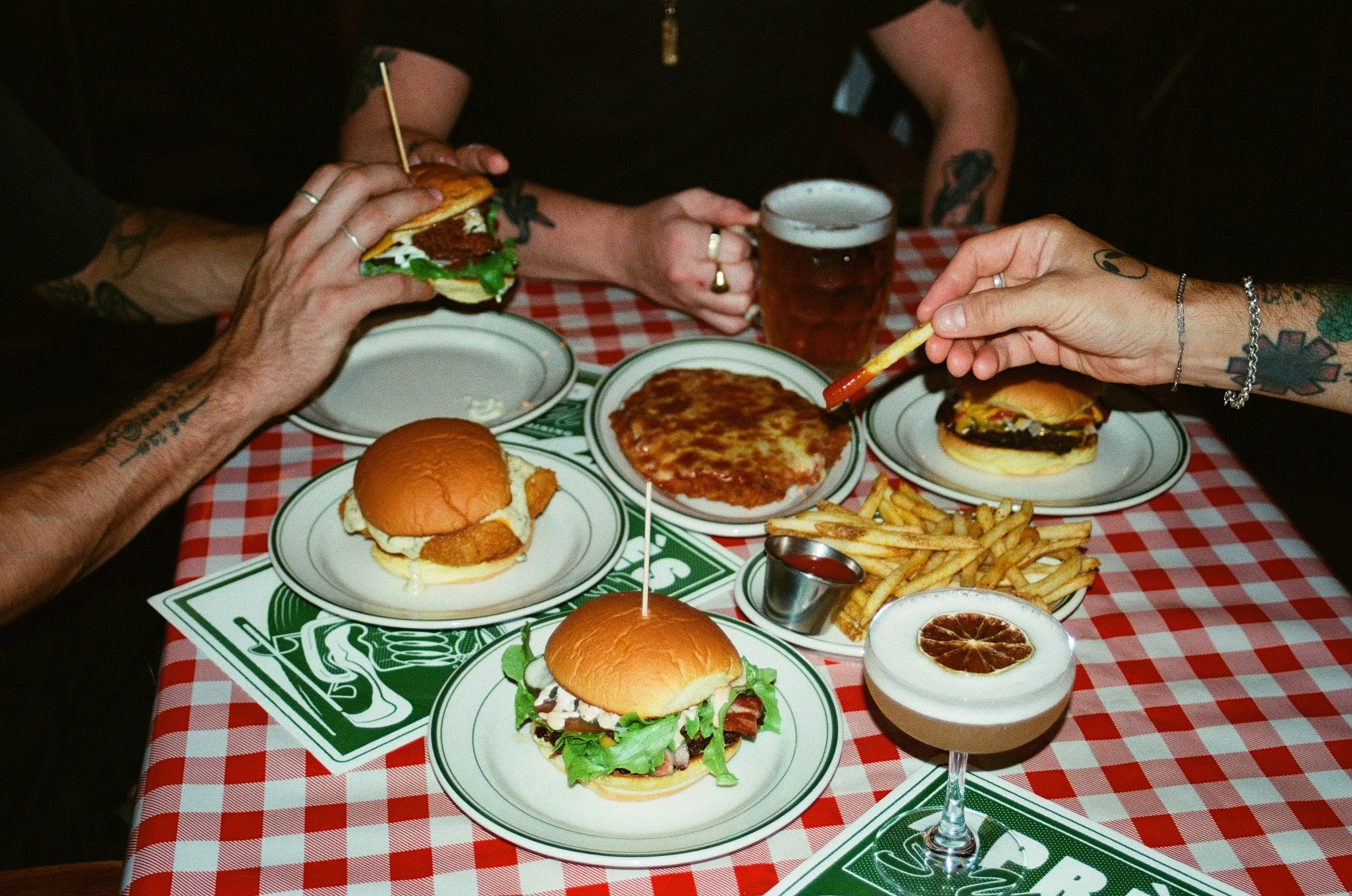A group of people enjoying a meal at a restaurant table covered with a red and white checkered tablecloth. The table has various foods including cheeseburgers, fries, pizza, and a drink with a lemon slice. Apres Surf Kitchen and bar