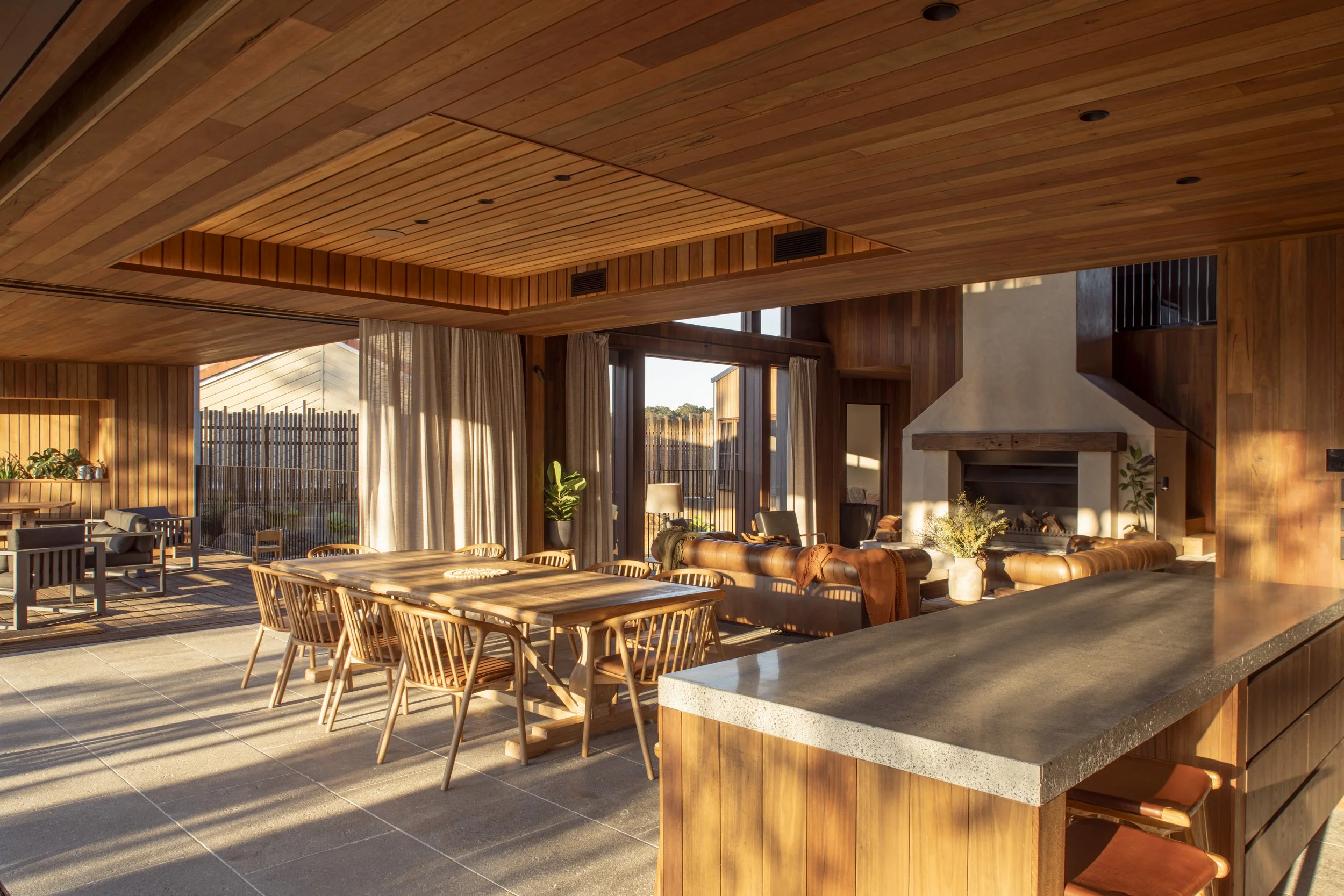 Open indoor living area with wooden walls, ceiling, and furniture, featuring a dining table, chairs, a sitting area with leather sofas, a fireplace, and an outdoor patio with additional seating. Currumbin Architecture Photography Joey Bailey