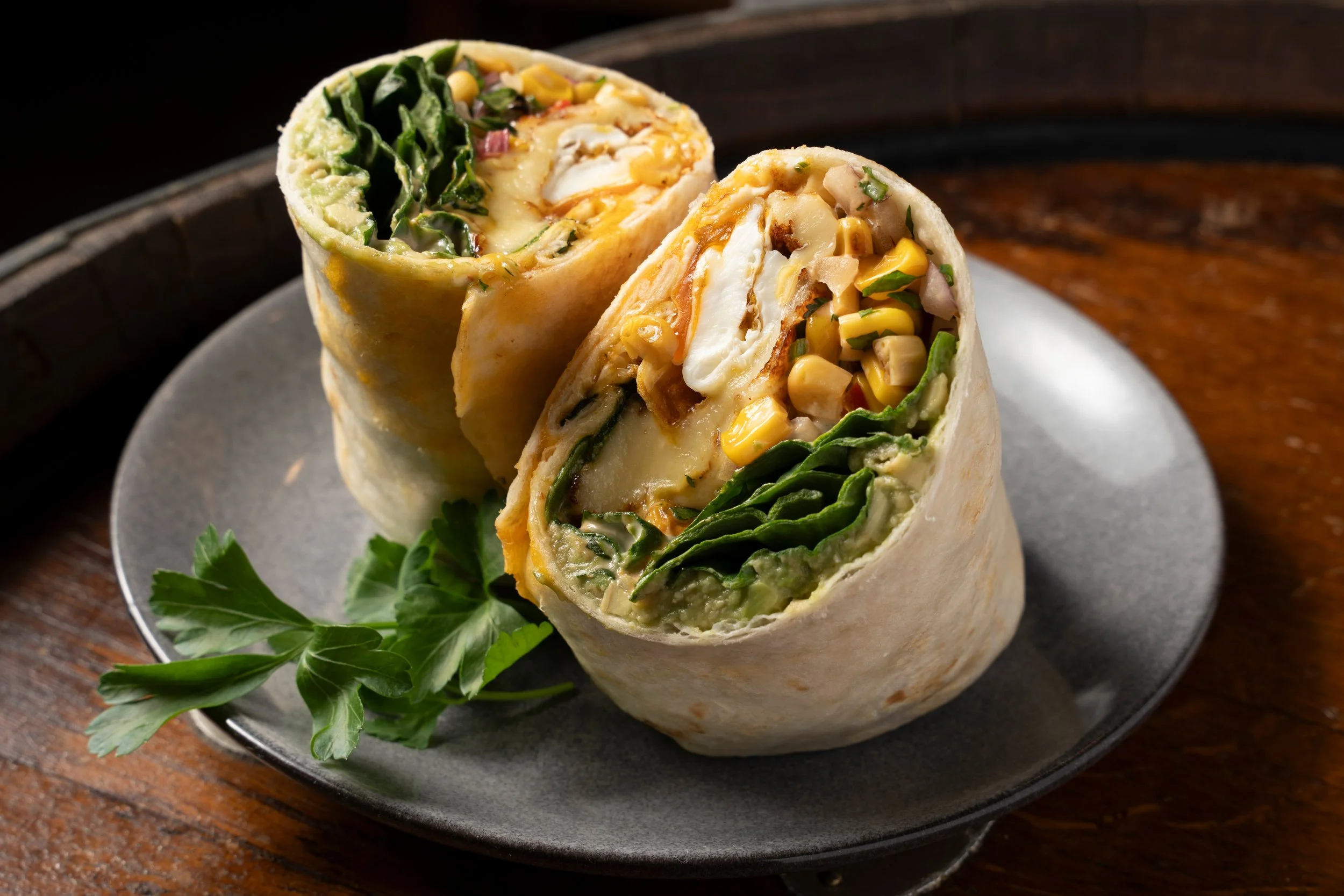 Two vegetable wraps sliced in half, showing fillings like lettuce, corn, cheese, and other vegetables, on a black plate with a sprig of parsley. Gold Coast food photography by Joey Bailey