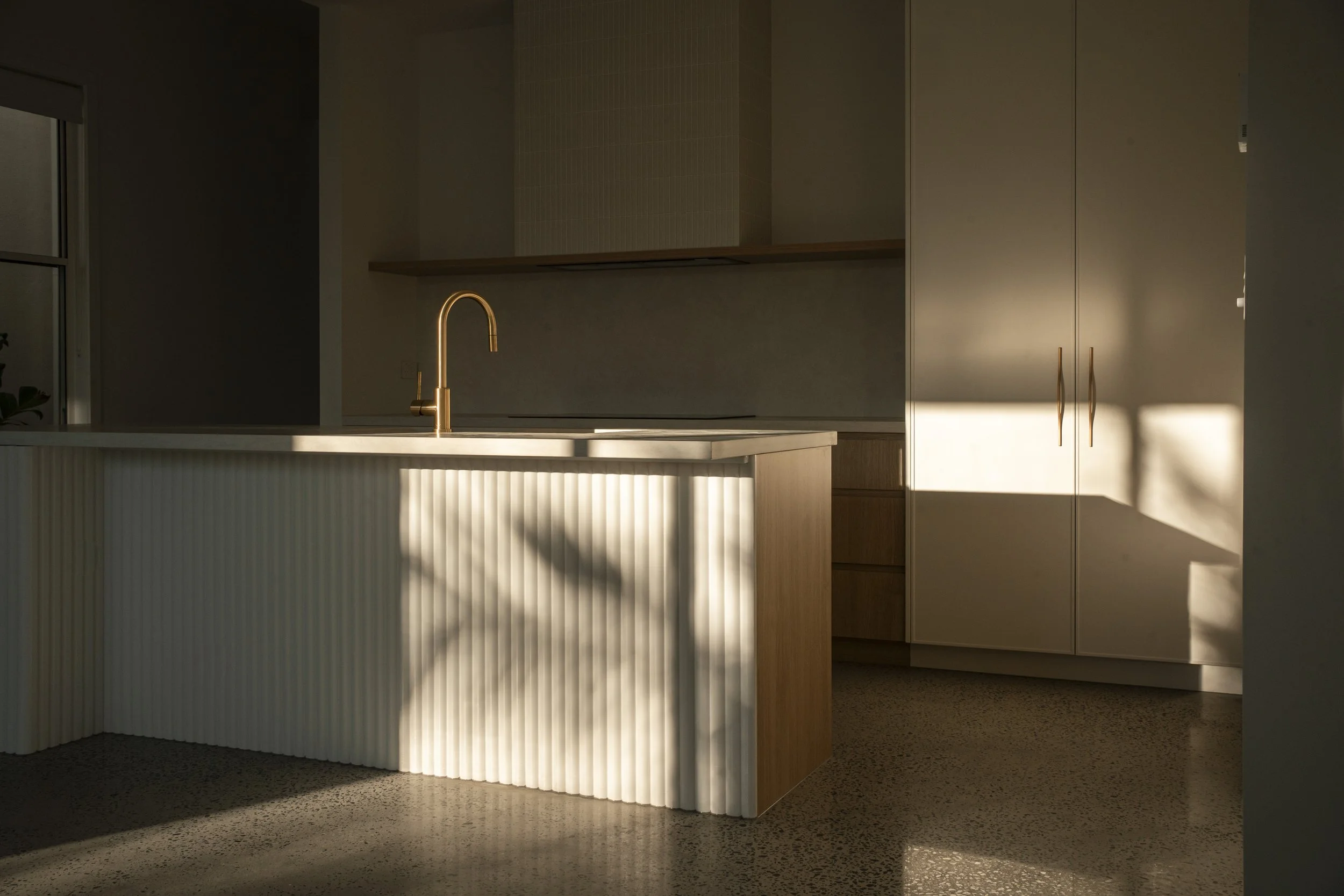 Modern kitchen with beige cabinets, a white kitchen island with vertical ridges, a gold faucet, and sunlight casting shadows on the wall and floor. Architectural photography by Joey Bailey, Gold Coast