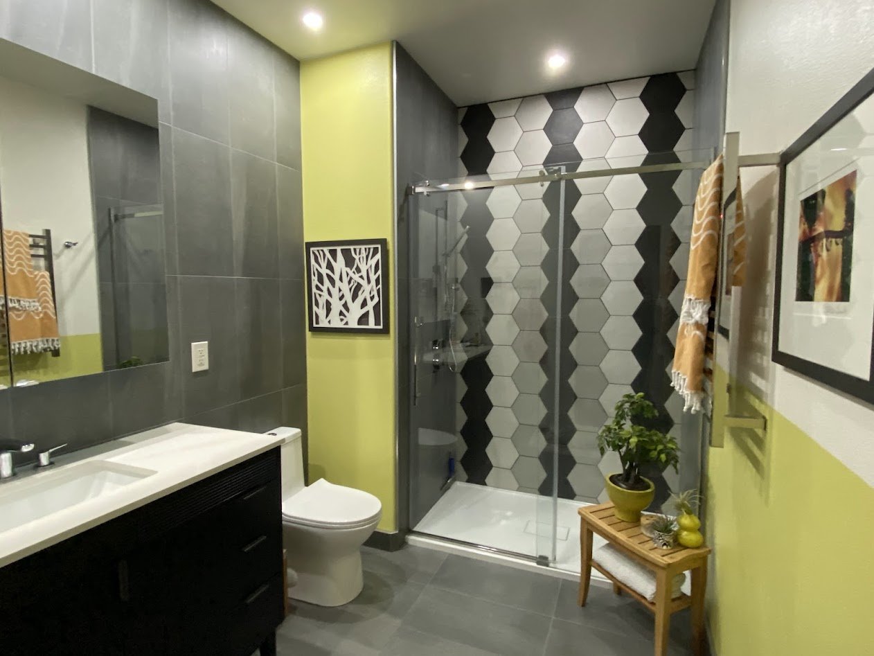 Modern bathroom with gray tile walls, a glass shower with hexagon tiles, a white toilet, a black vanity with a white sink, a mirror, yellow and gray walls, framed artwork, and a small wooden table holding a potted plant and decor items.