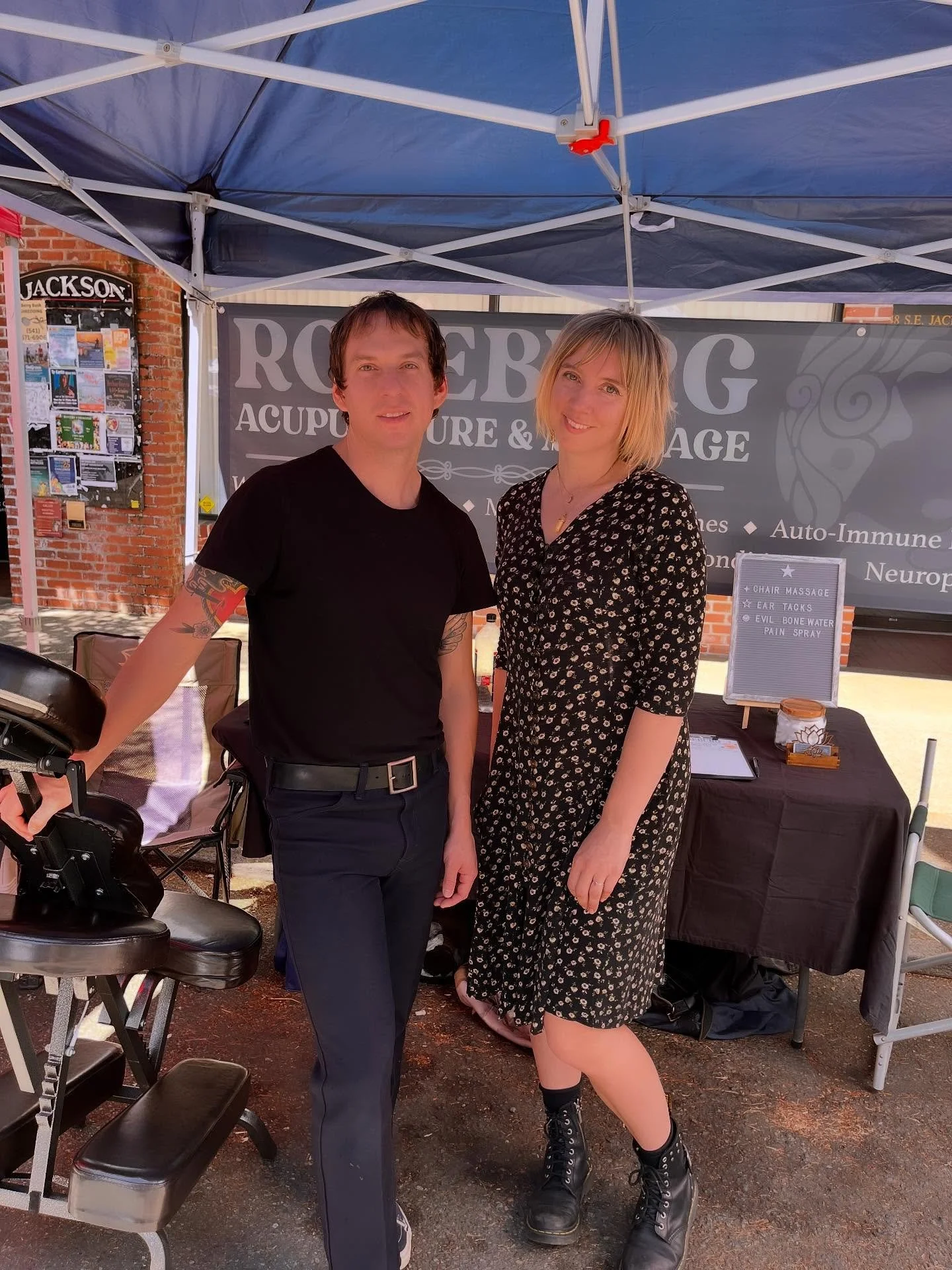 Two people standing under a blue canopy, smiling. Behind them is a sign that reads 'ROSEBLOOM ACUPUNCTURE & MASSAGE'. One person is a man with dark hair in a black t-shirt and dark pants, and the other is a woman with blonde hair in a black dress with a floral pattern, black boots, and an anklet.