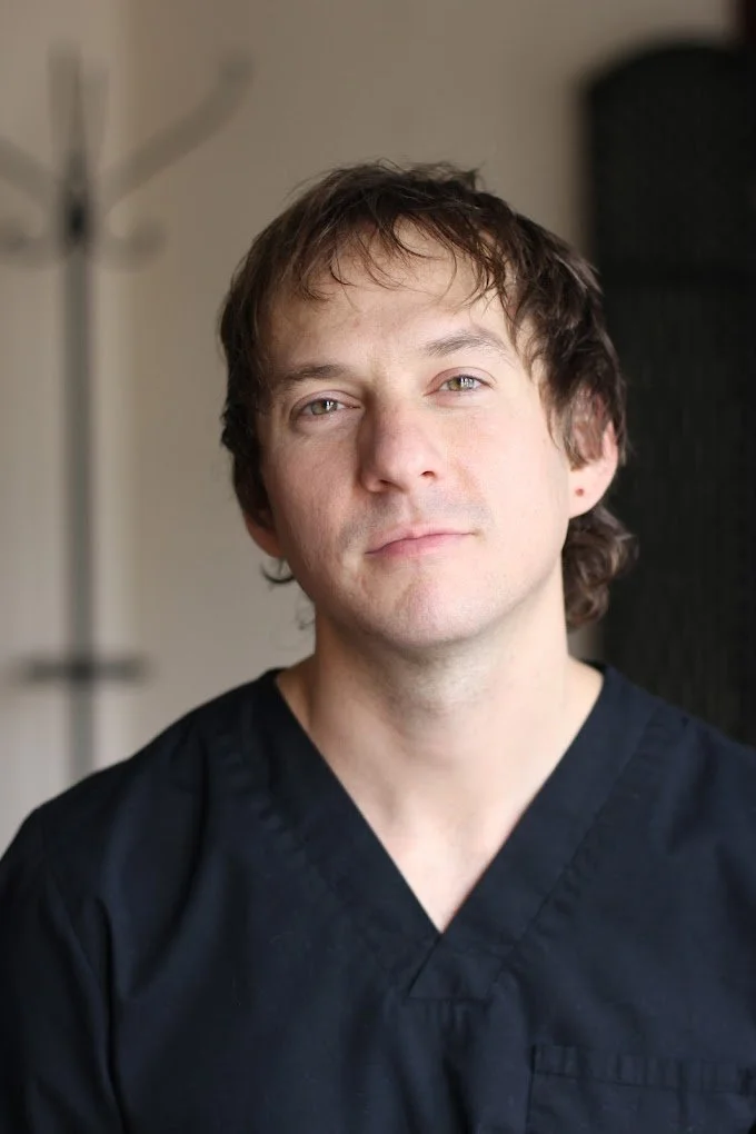 A man in black medical scrubs with light brown, slightly curly hair looking at the camera, in a setting with a coat rack in the background.