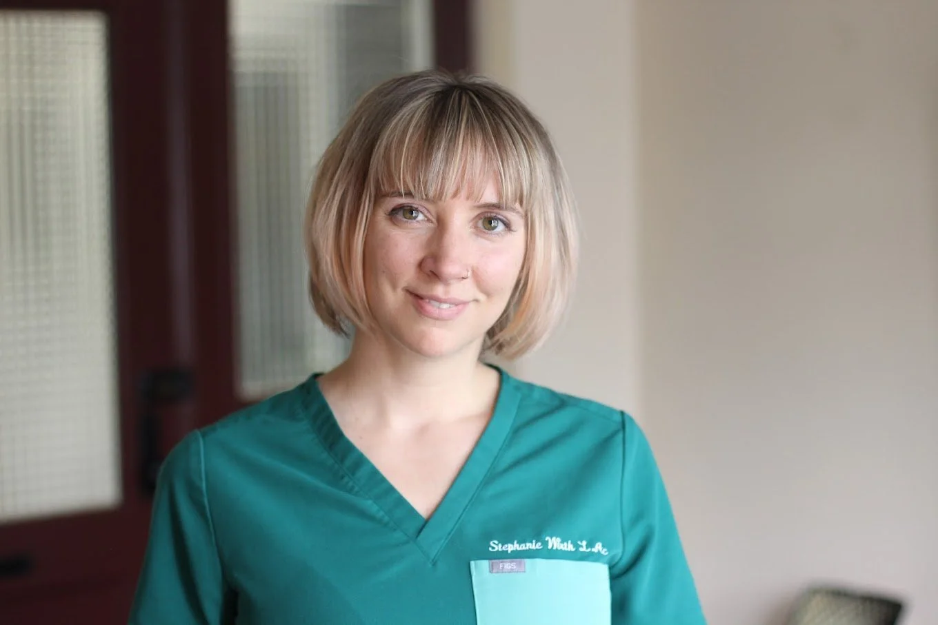 A woman with short blonde hair wearing green scrubs with a name tag that says "Stephanie Wuth L. A." standing in a room with blurred background.