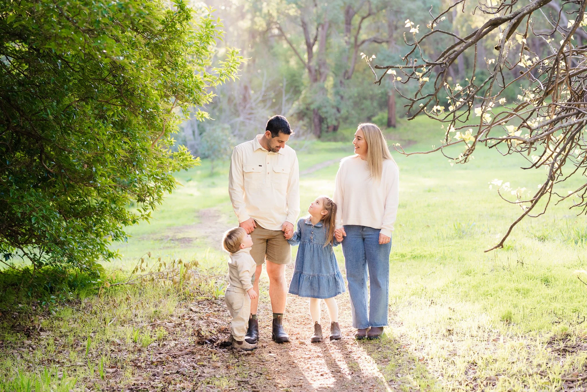 A family of four walking together on a sunlit trail in the Perth Hills surrounded by trees, holding hands and smiling at each other.