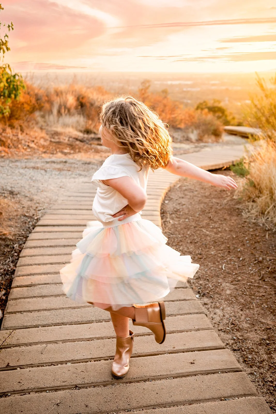 A young girl twirling on a wooden pathway during a sunset, wearing a pastel rainbow tutu skirt and tan boots, with her hair flowing and arms extended.